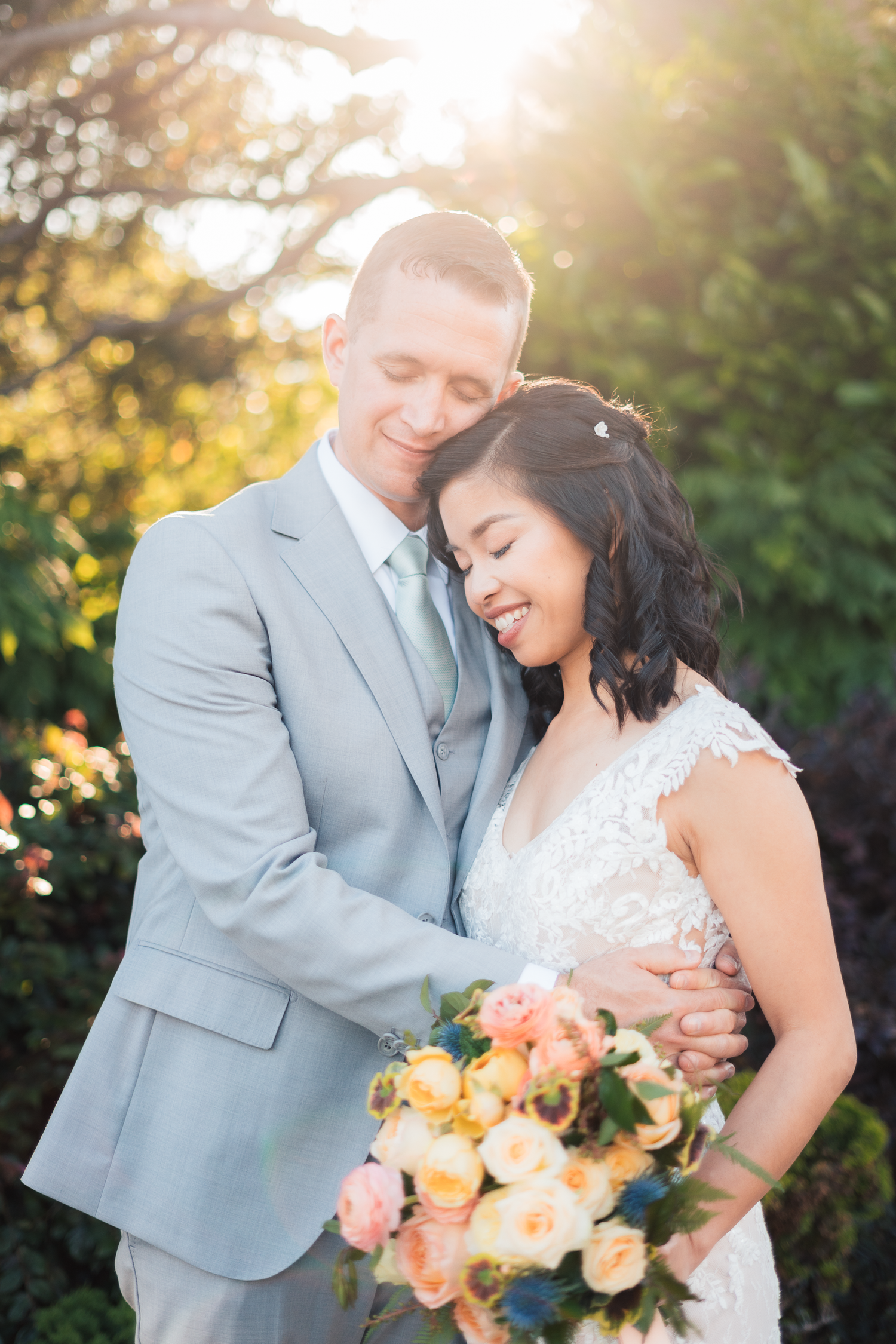Newlywed couple shares romantic moments in garden setting with peach floral bouquet at sunset.