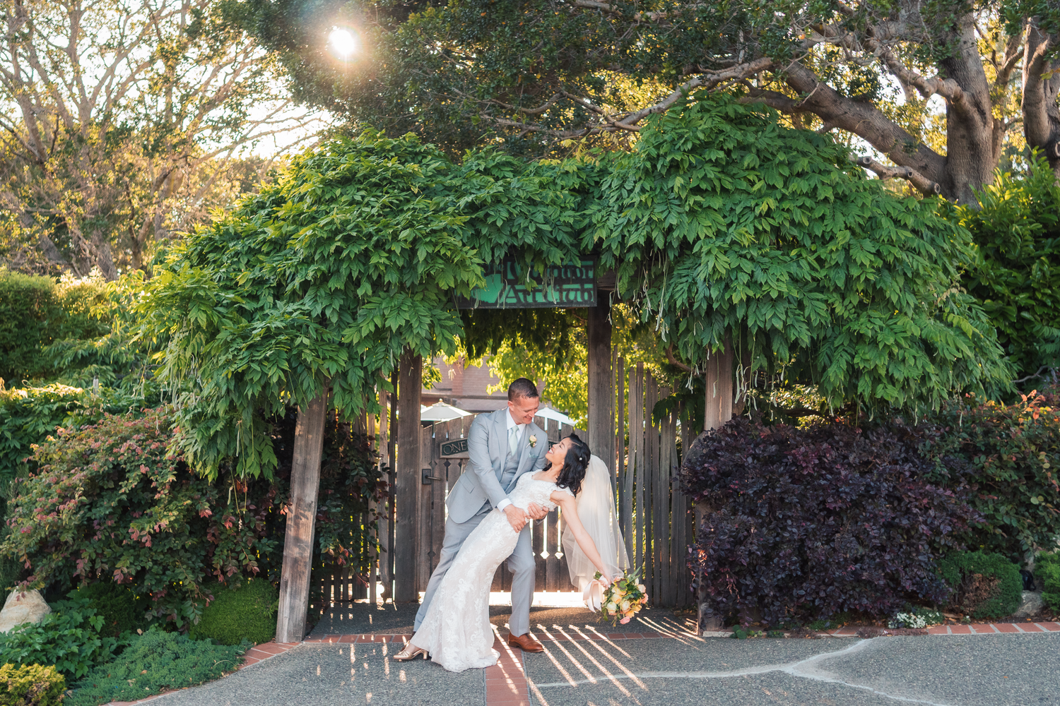 A person in a white dress twirls playfully in a sunlit garden path surrounded by lush greenery and trees.