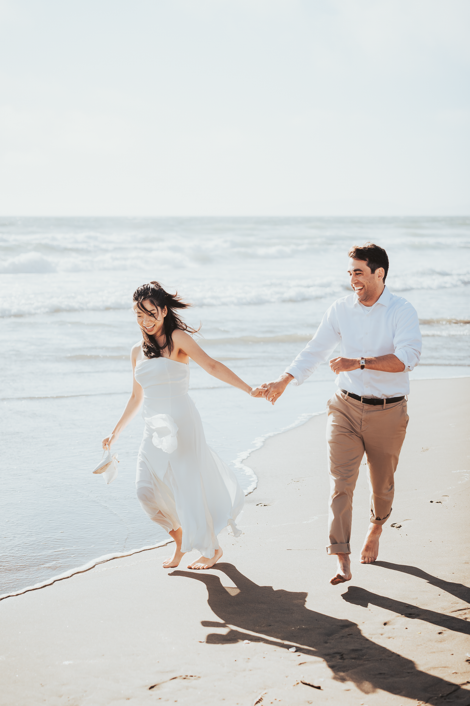 Couple runs playfully along beach shoreline in white and tan outfits at Ocean Beach in San Francisco.