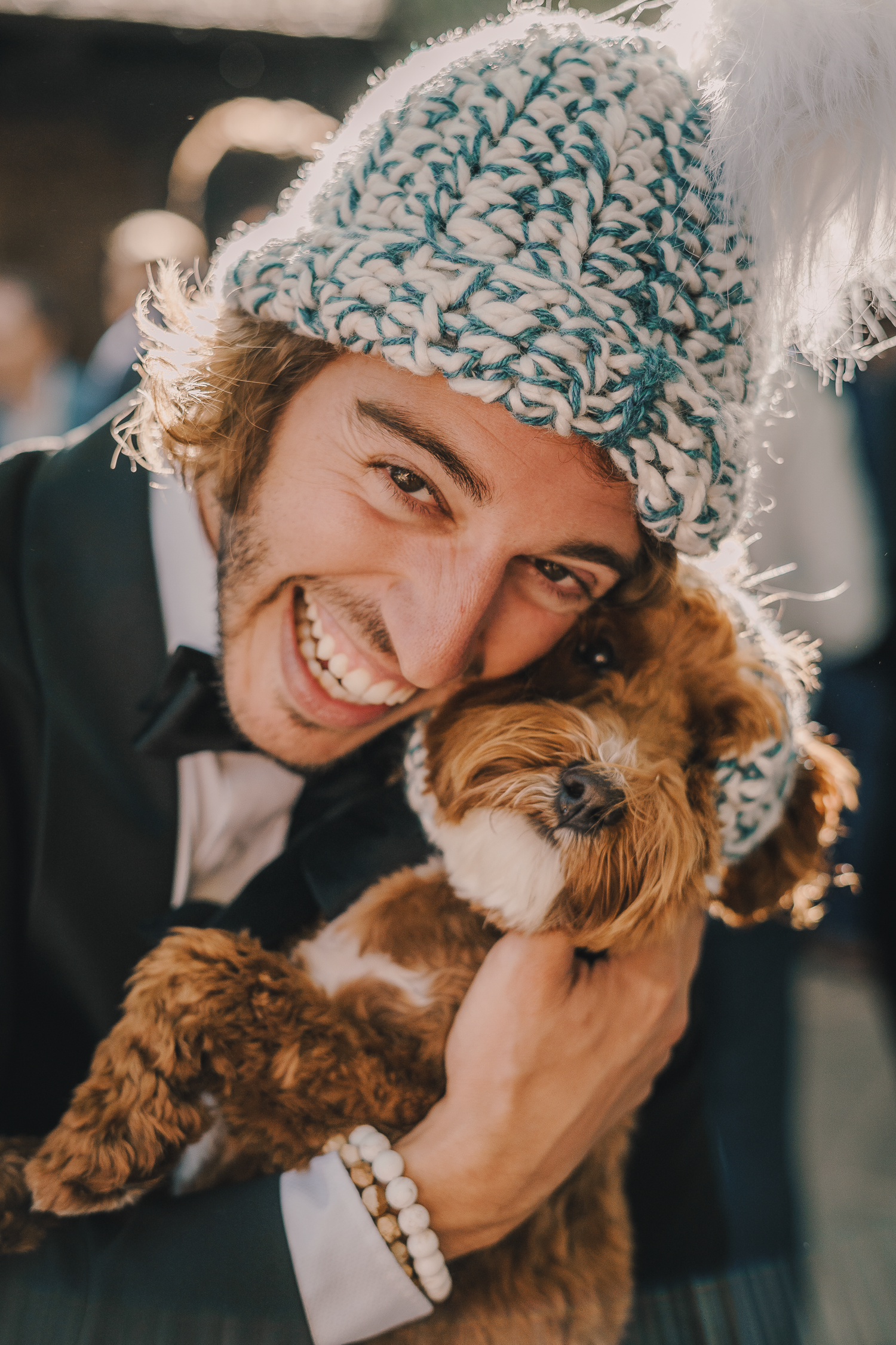 Groom in winter hat cuddling with small brown dog while wearing formal attire.