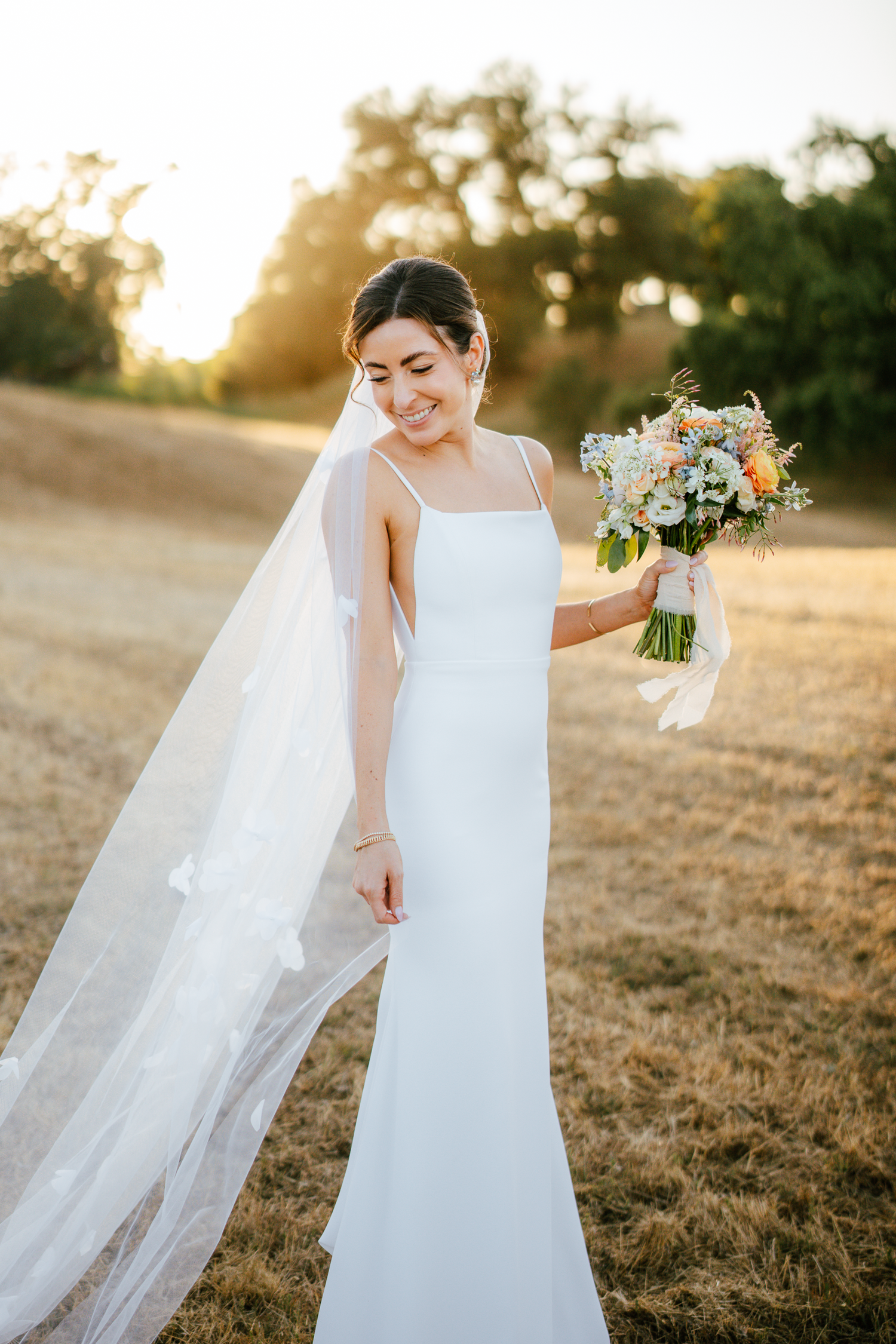 Bride in a simple elegant white dress holding a bouquet in a sunlit field at Harper's Rest in Healdsburg, California.