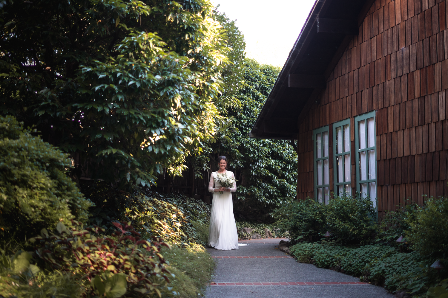 A couple in wedding attire stands together near a rustic wooden barn surrounded by lush greenery and natural light.