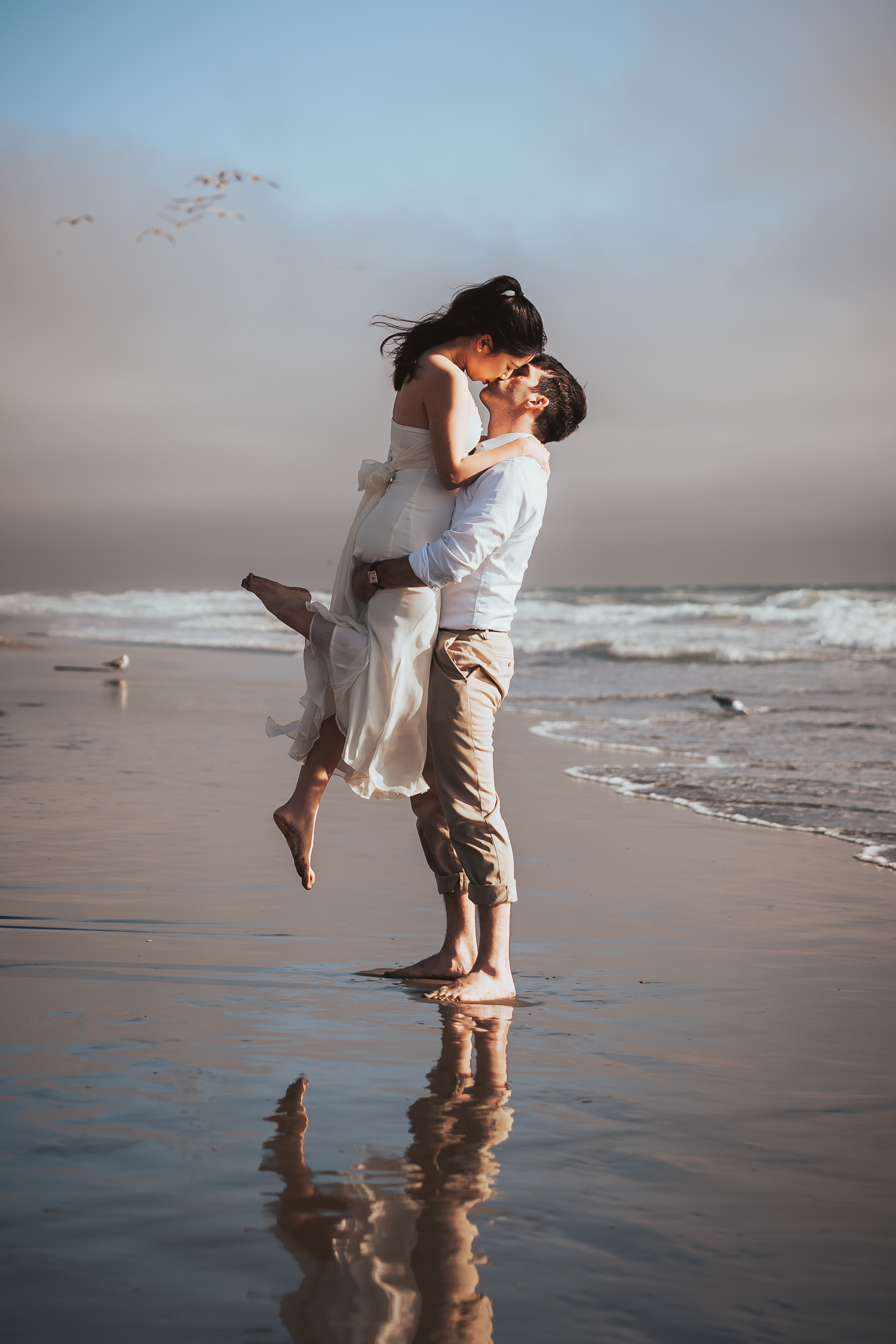 A romantic couple dances and embraces on a misty beach at sunset with waves crashing in the background at Ocean Beach in San Francisco.