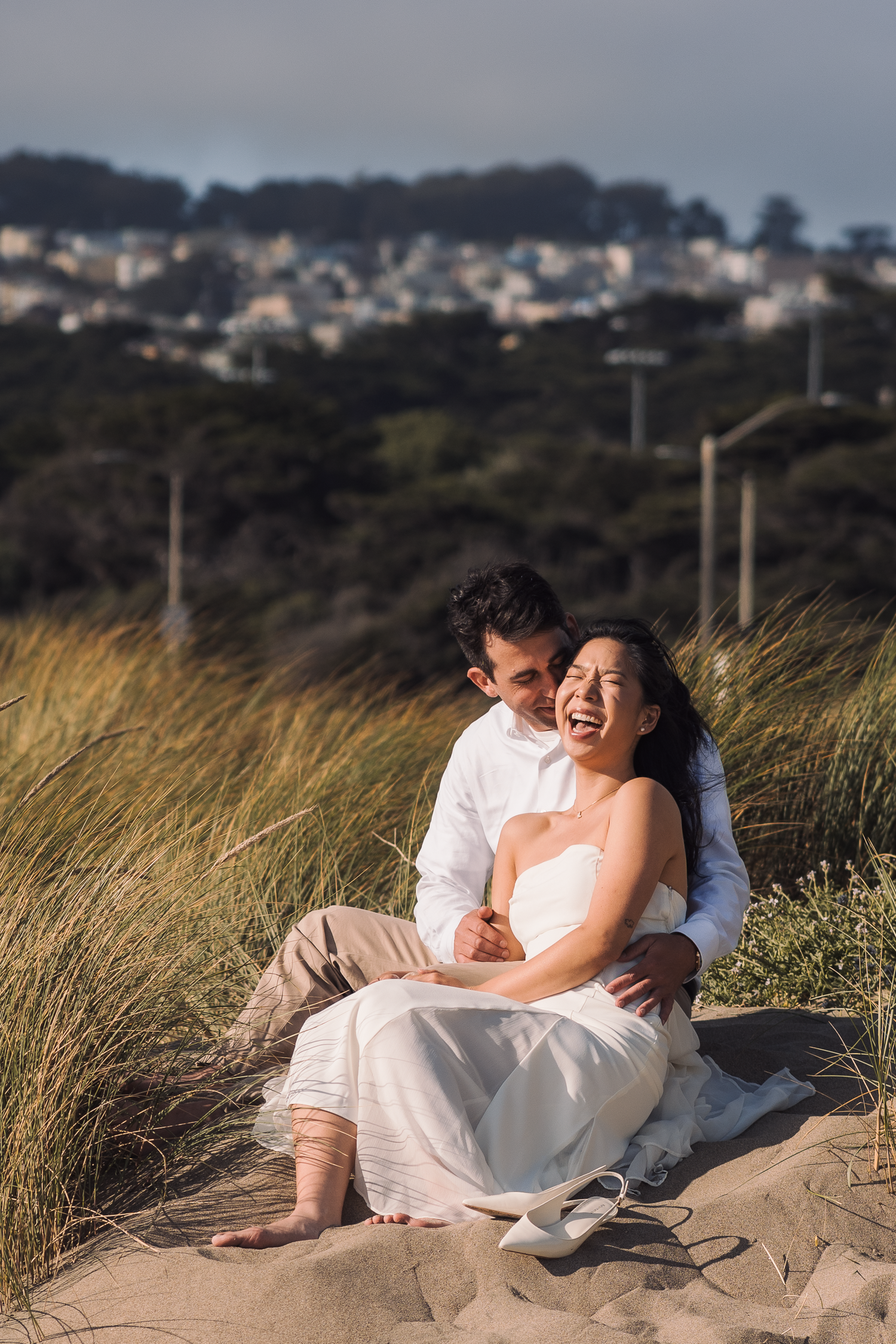 Couple cuddles on sand dunes with coastal town visible in background at Ocean Beach in San Francisco.