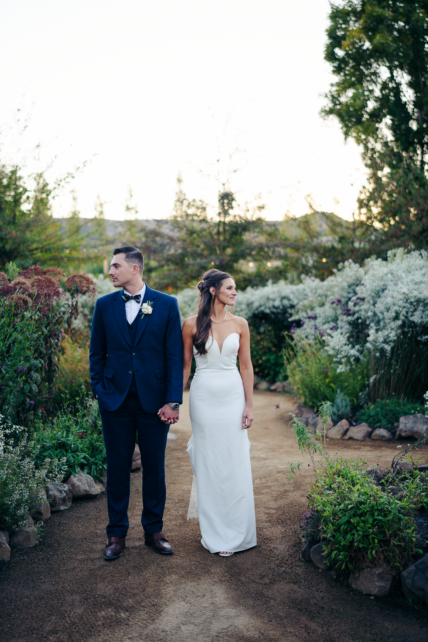 Newlyweds stroll through a garden path surrounded by blooming white flowers at sunset at Cornerstone Sonoma.