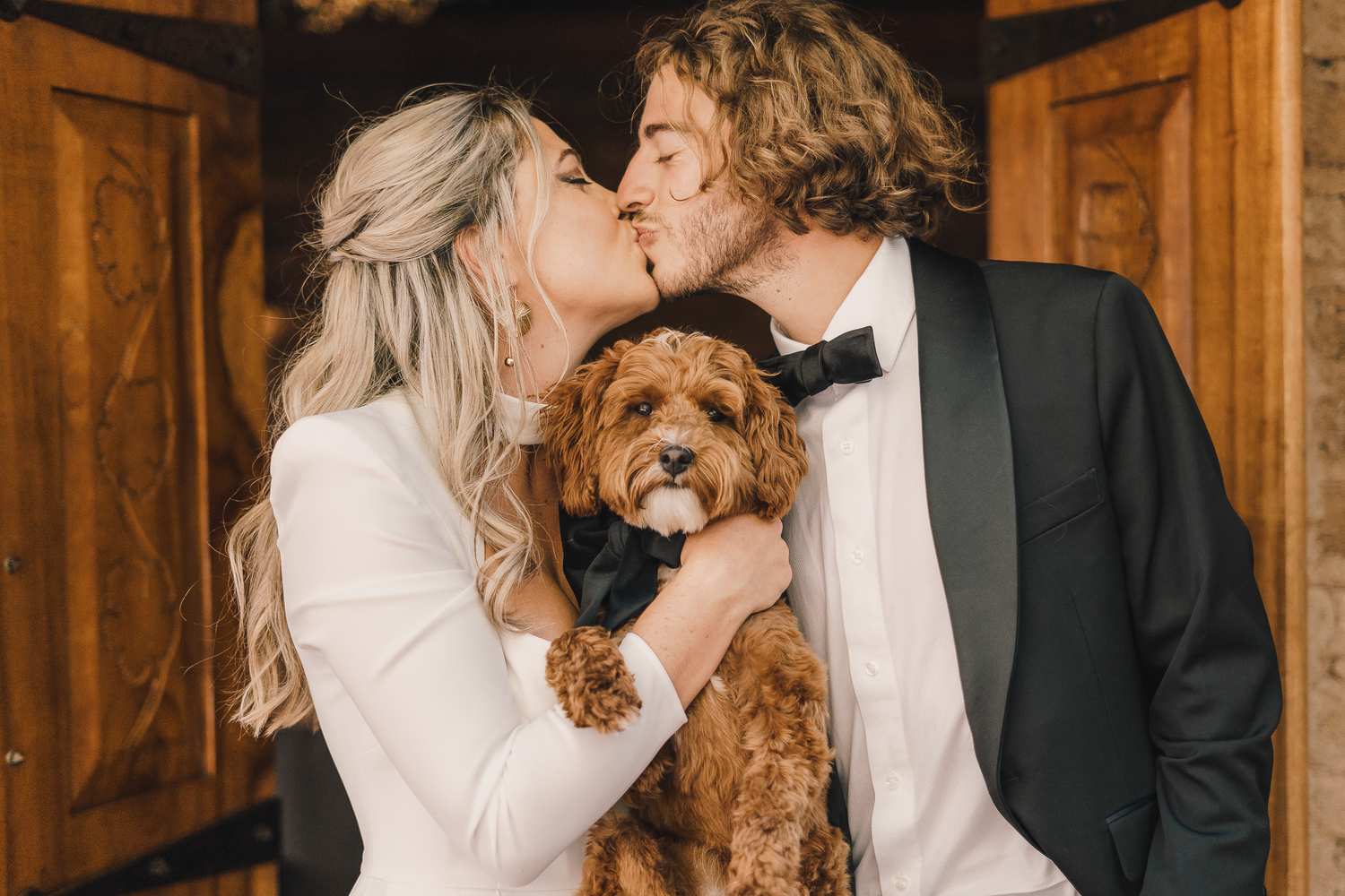 Bride and groom shares romantic kisses in a rustic wooden venue with their dog.