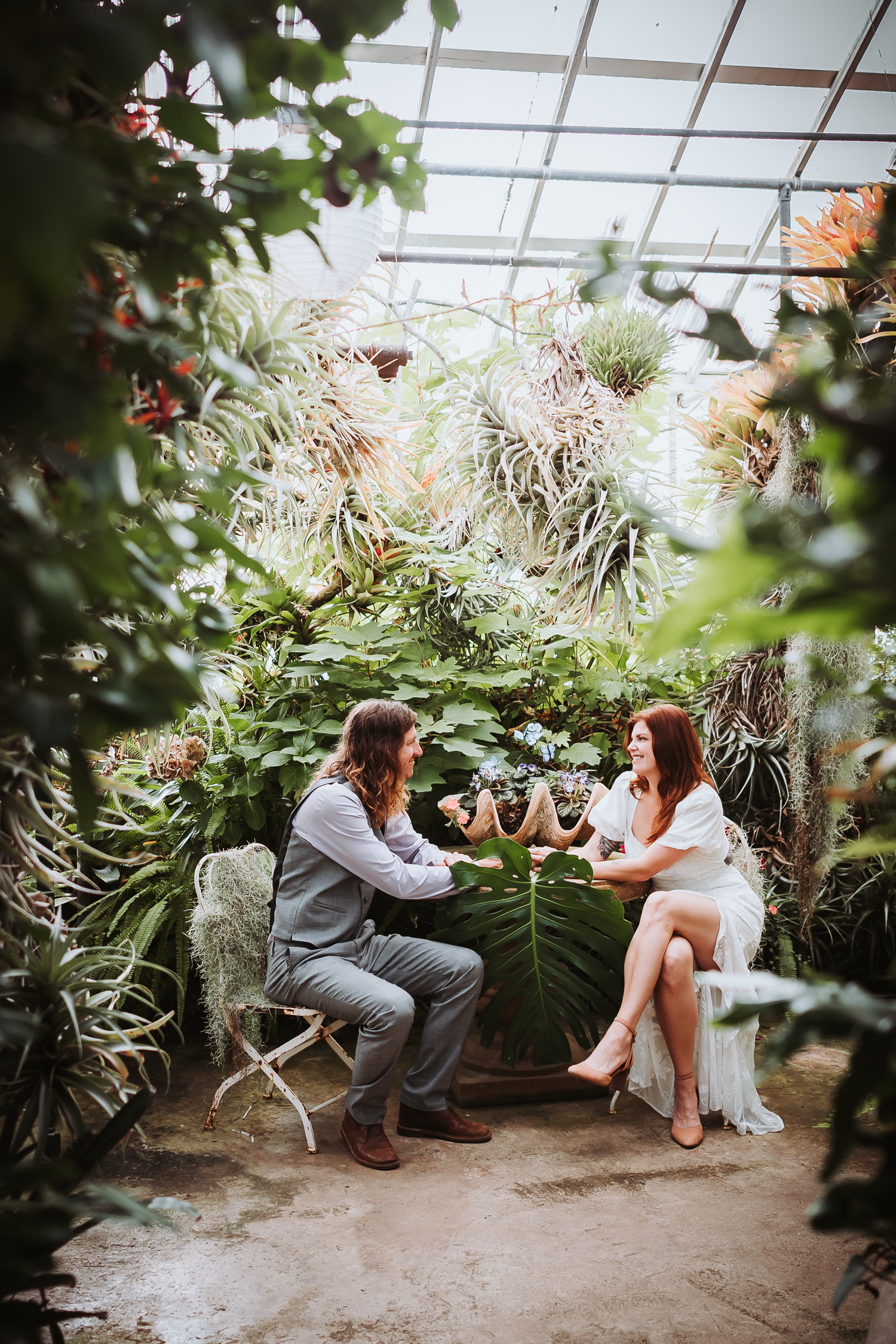 Couple sitting together in a greenhouse surrounded by tropical plants and hanging air plants at Shelldance Orchid Farm in Pacifica.