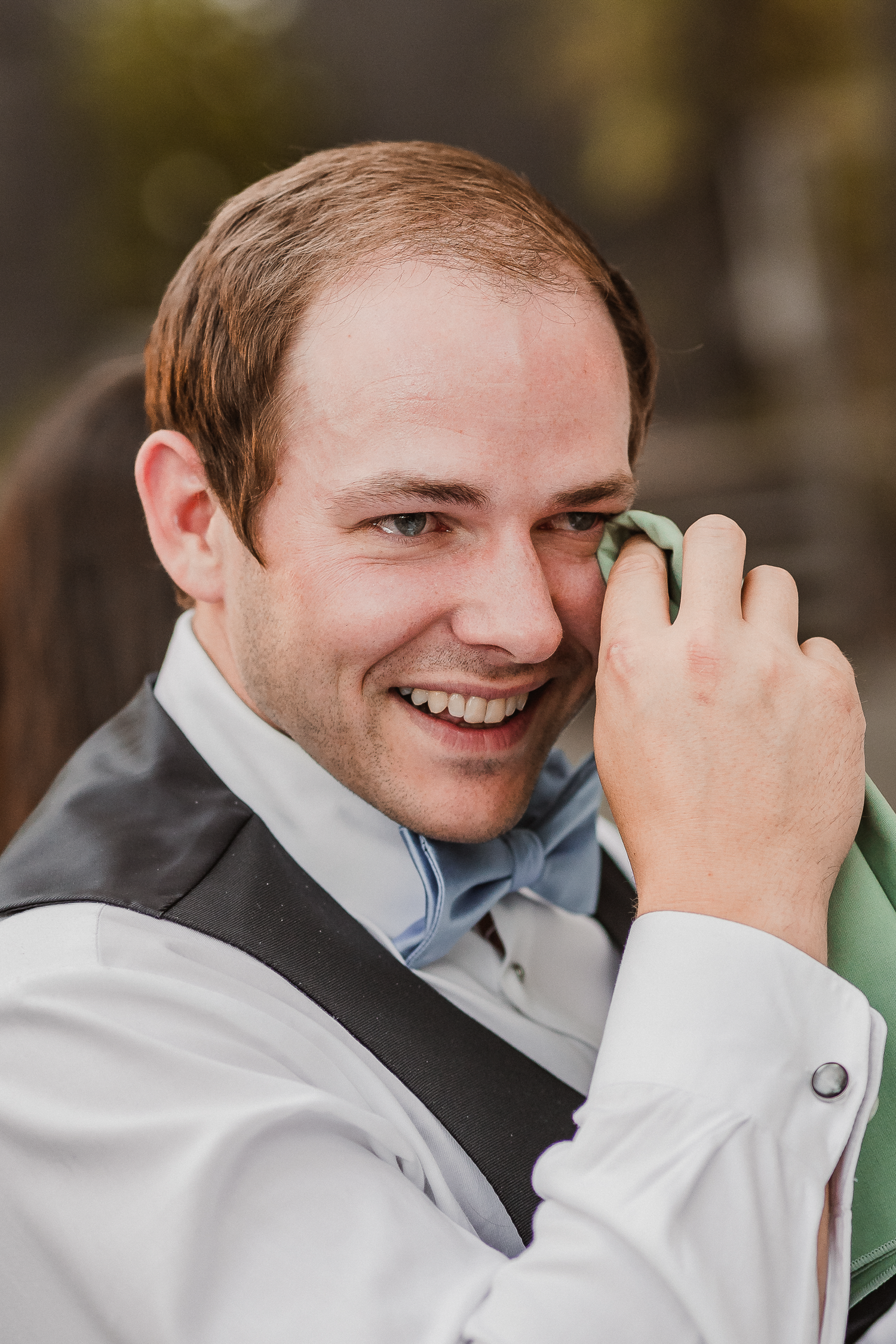 Groom in formal attire wiping eye while smiling at camera.