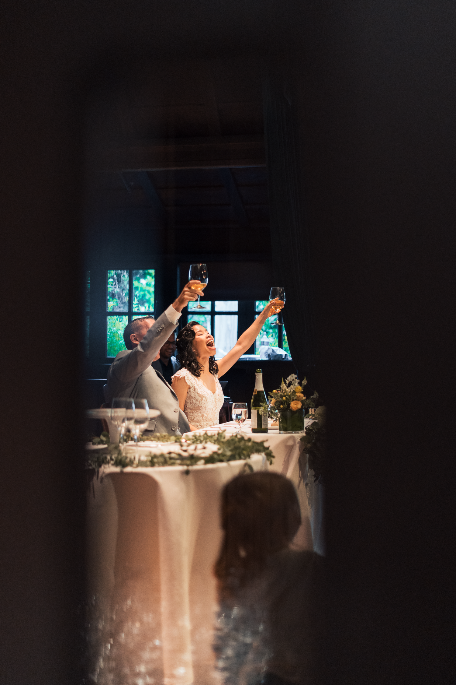 A couple raises their glasses in celebration at a dimly lit wedding reception table decorated with candles and flowers.