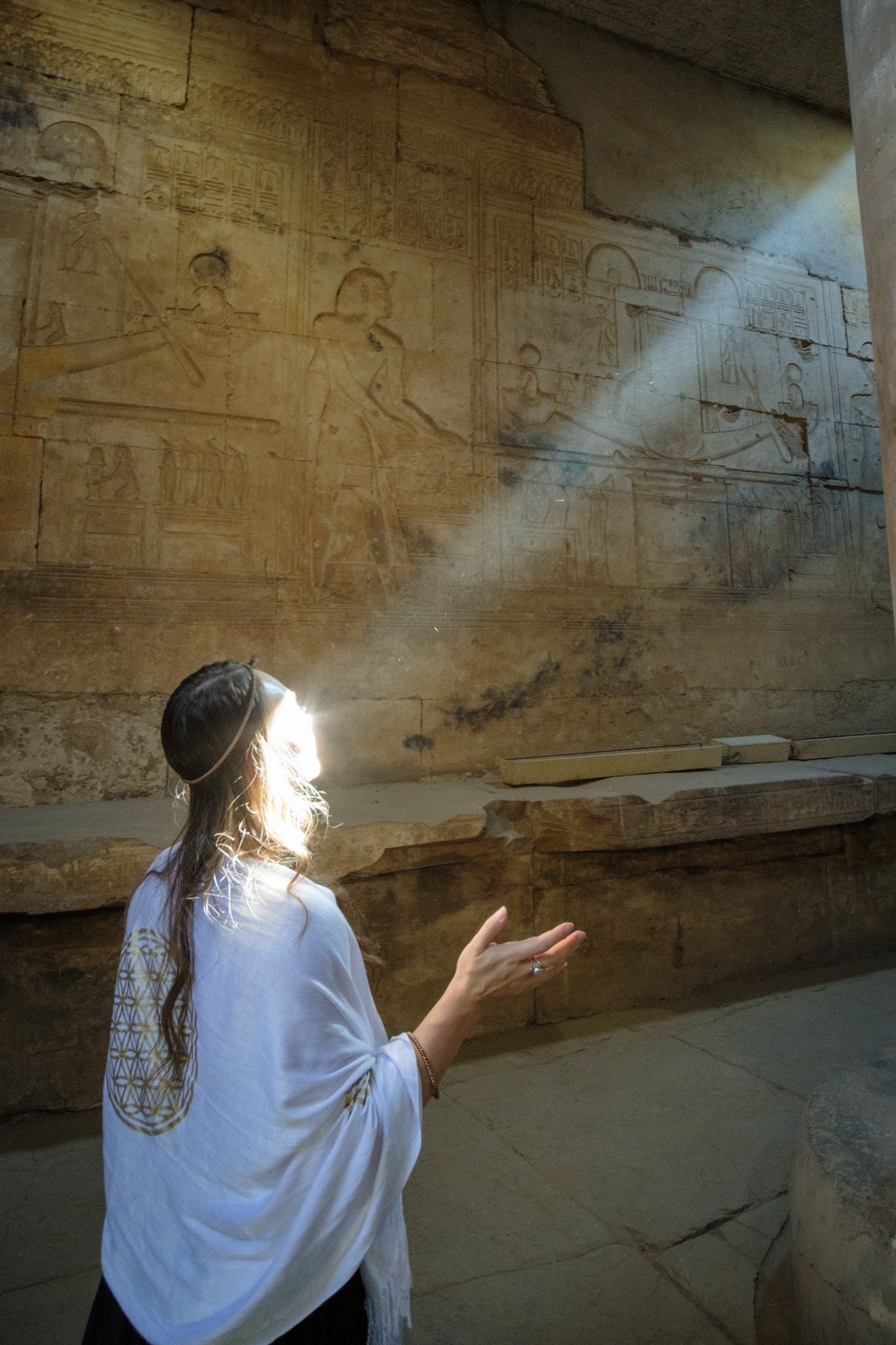 A person in white clothing stands in a beam of sunlight while looking up at ancient Egyptian hieroglyphic carvings on a temple wall.