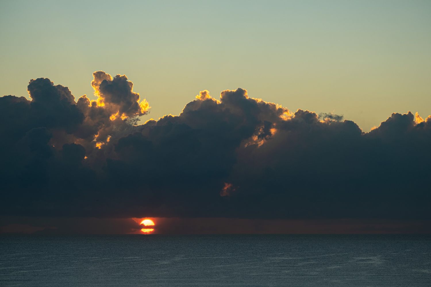 Dramatic sunset over ocean with sun peeking through dark cloud formations.
