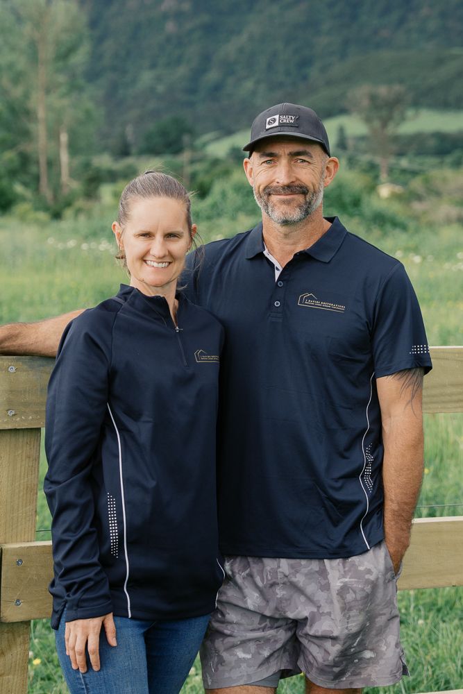Two people in matching navy polo shirts standing by wooden fence in outdoor setting.