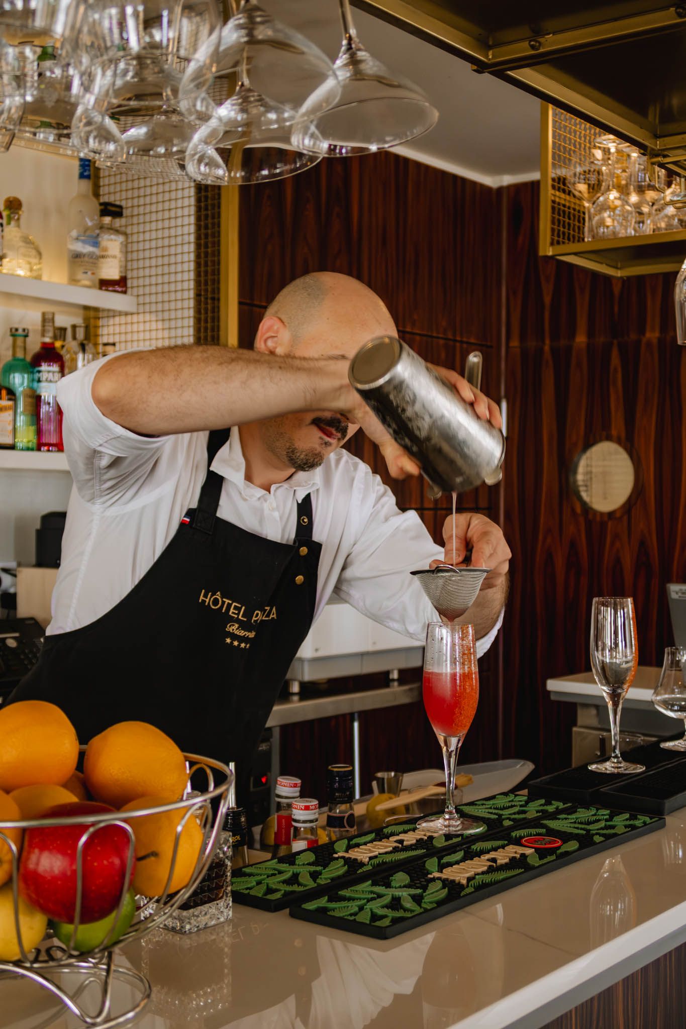 A bartender in a black apron carefully strains a cocktail into a glass while fresh citrus fruits sit nearby at an elegant bar.