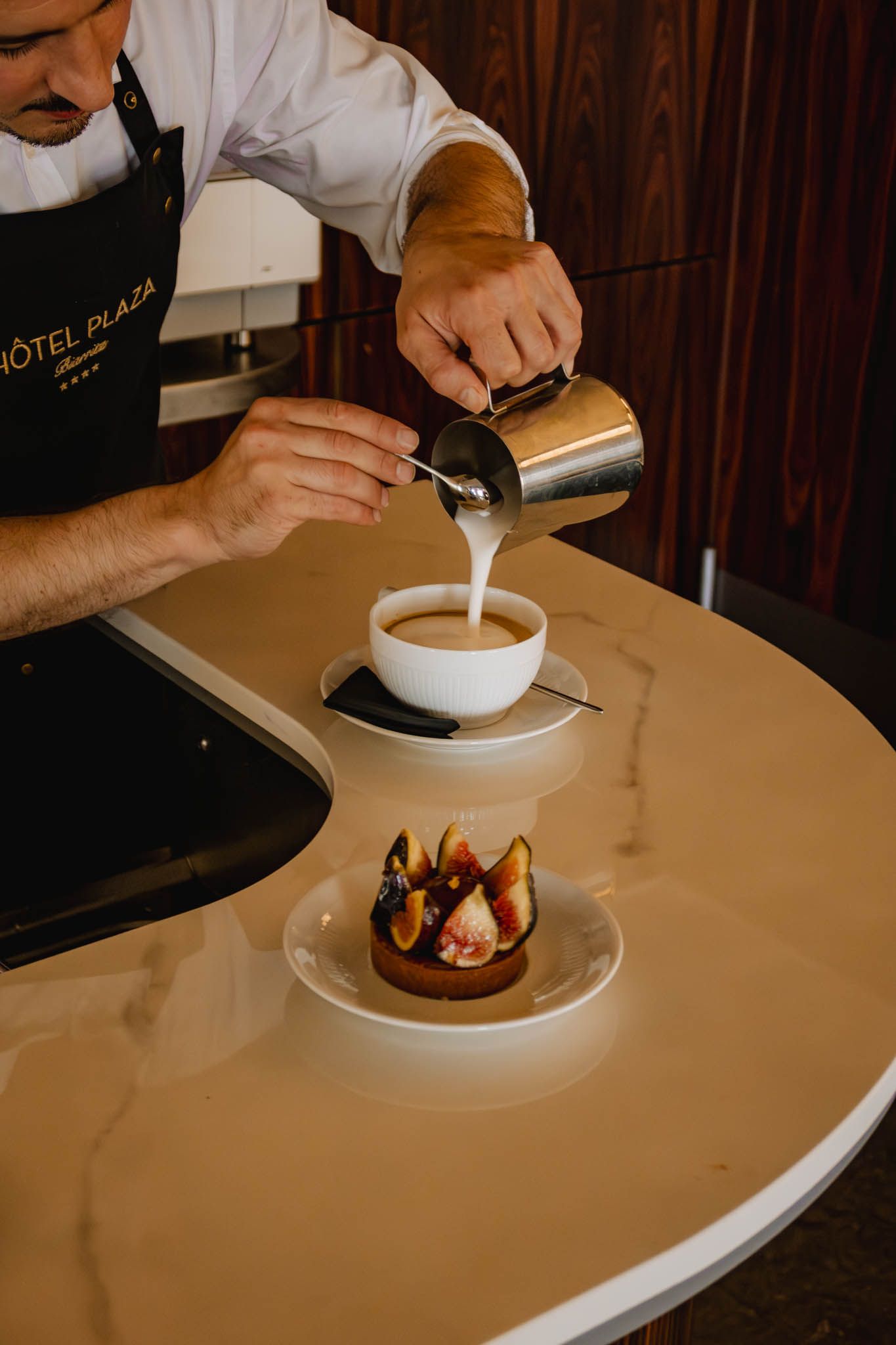 A barista pours steamed milk into a coffee cup next to an elegant dessert plate at a hotel cafe counter.