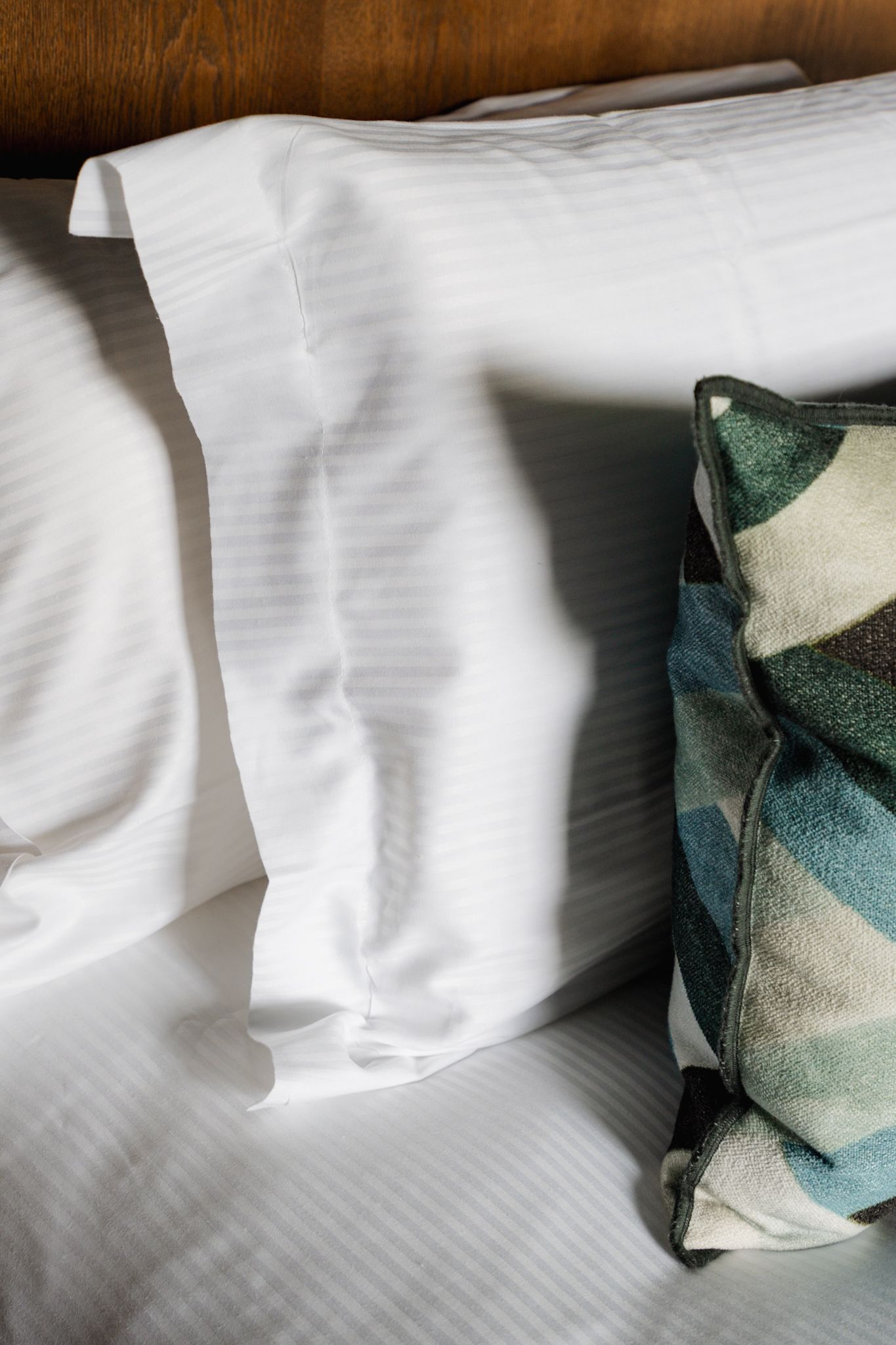 White cotton bed pillowcase with striped texture next to decorative geometric patterned cushion in green and blue tones.