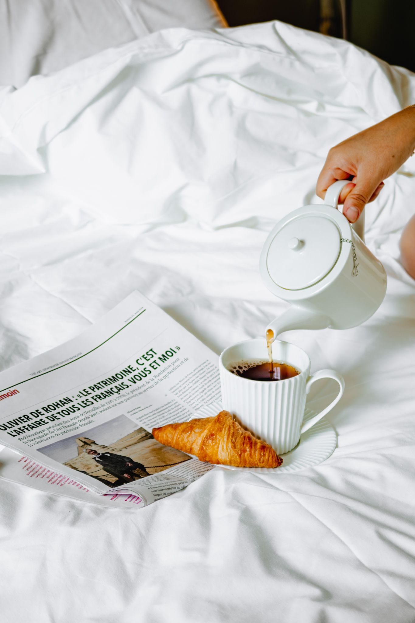 A cozy breakfast scene with a croissant, coffee cup, and newspaper on white bedding.