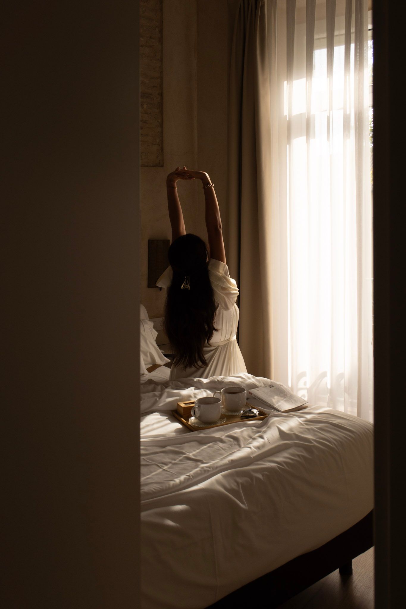 Silhouette of someone stretching in bed beside window with soft morning light filtering through sheer curtains.