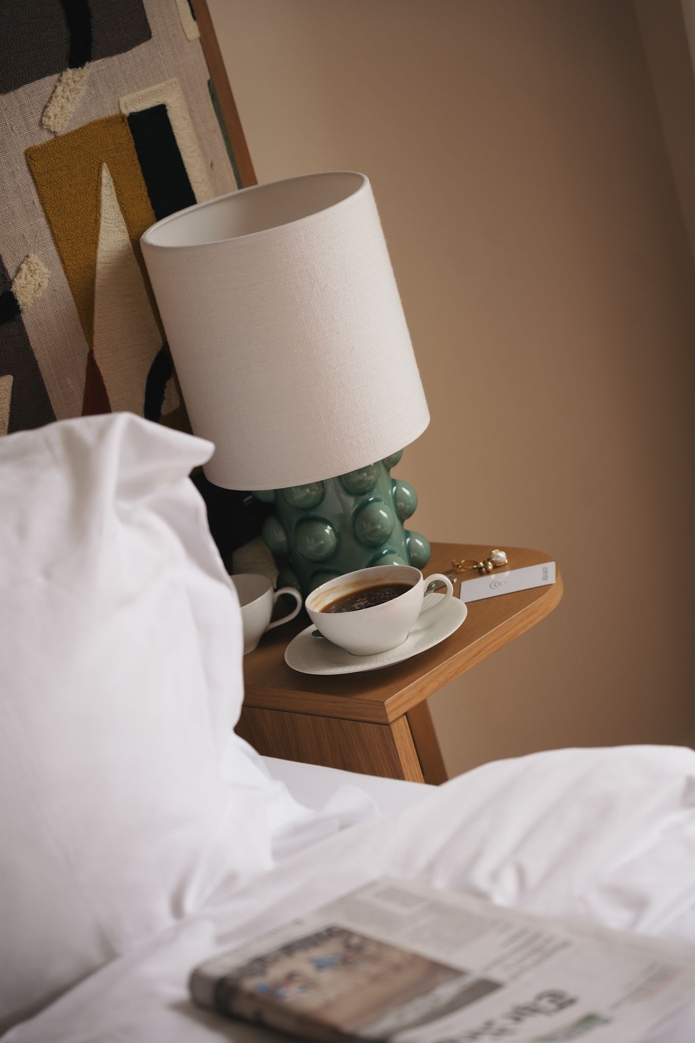 Reading book in bed with a white teacup on wooden tray, lamp with white shade casting warm light in bedroom.