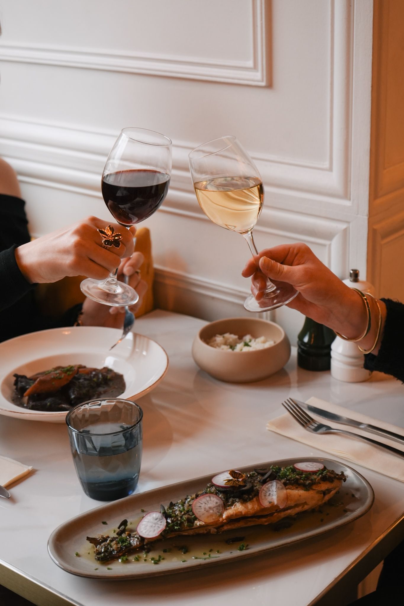 Two wine glasses clink together over a table set with elegant plates of food and a small bowl of appetizers.