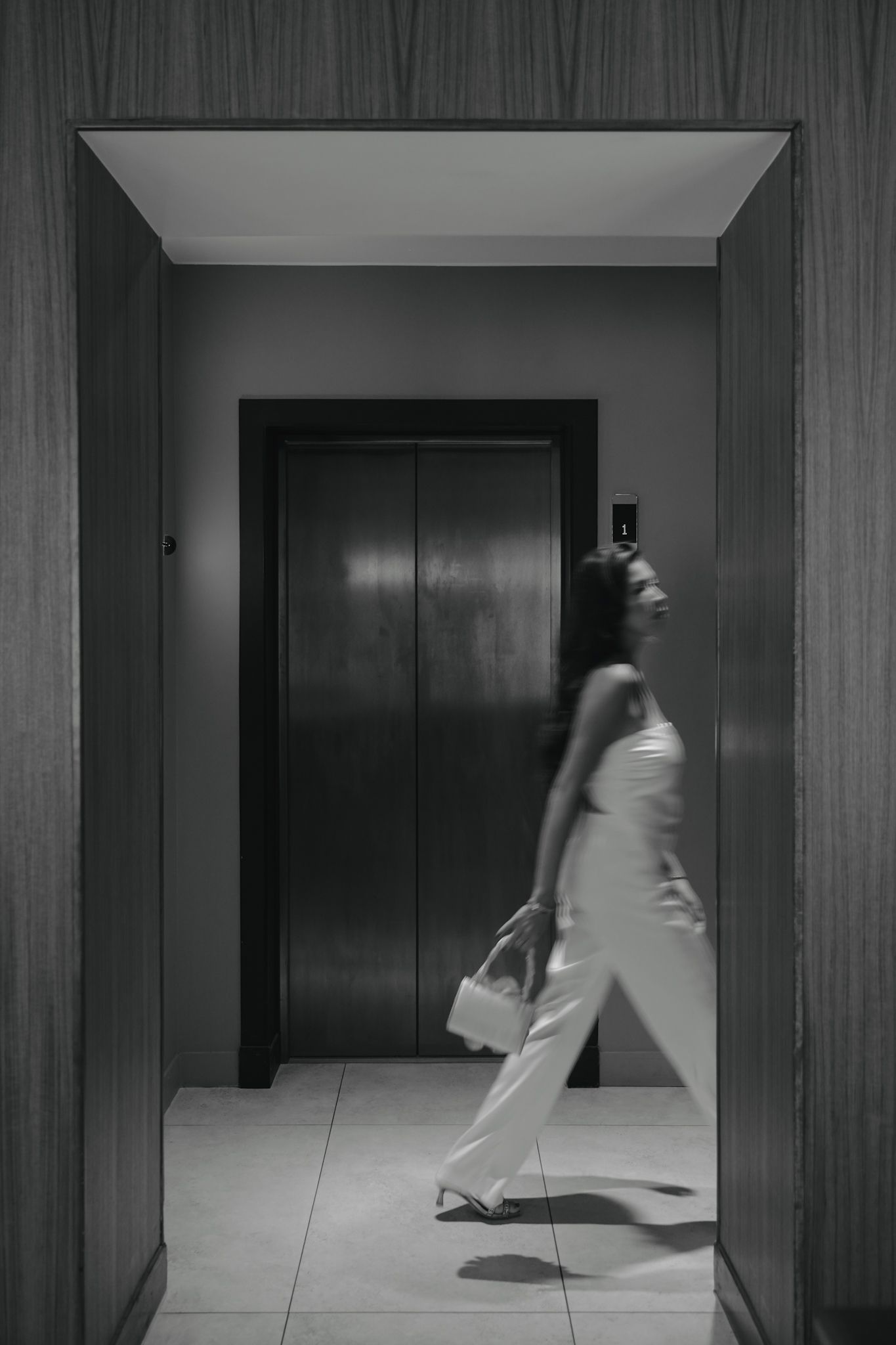 Black and white photo of someone in white attire walking past an elevator in a modern hallway interior.