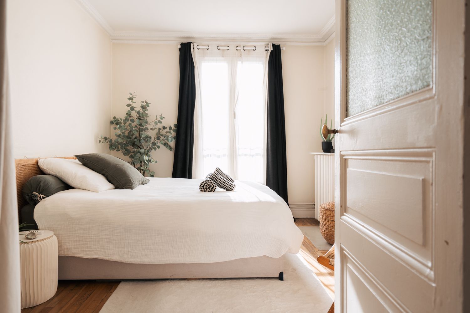 Minimalist bedroom interior with white bedding, black curtains, potted plant, and natural light streaming through window.