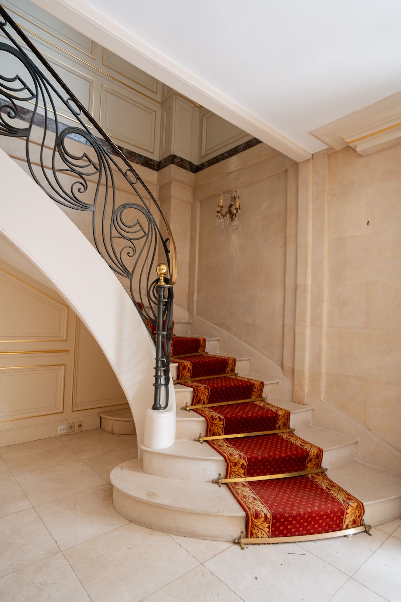 Elegant curved marble staircase with ornate iron railing and red carpeted steps in a classic interior space.