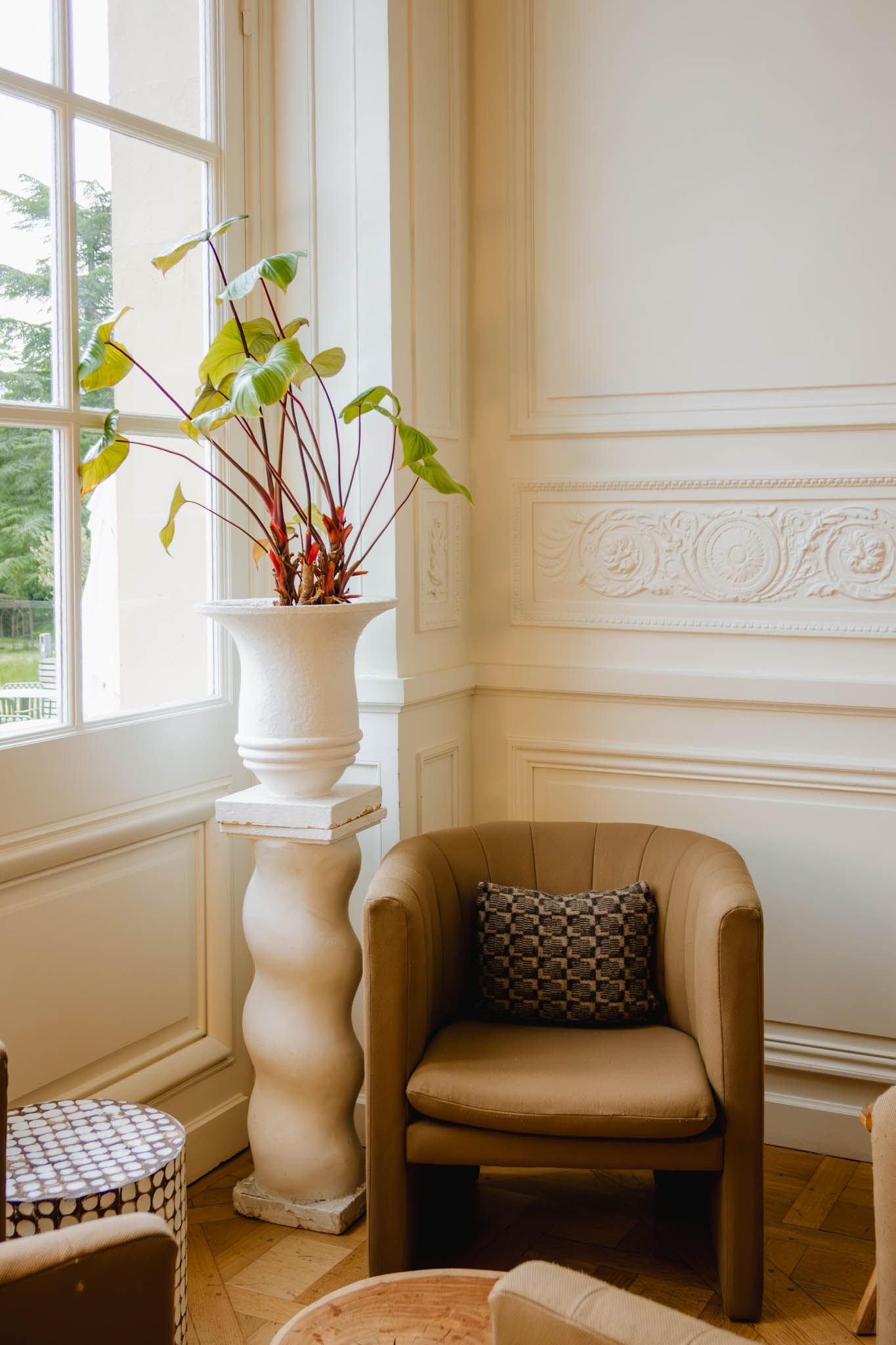 Modern sitting corner with tan barrel chair, decorative white pedestal, and potted plants near bright window.