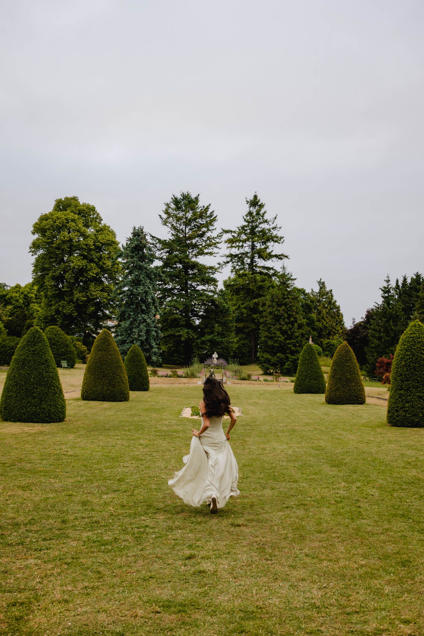 A figure in a flowing white dress dances through a manicured garden with sculptural topiary trees on a cloudy day.
