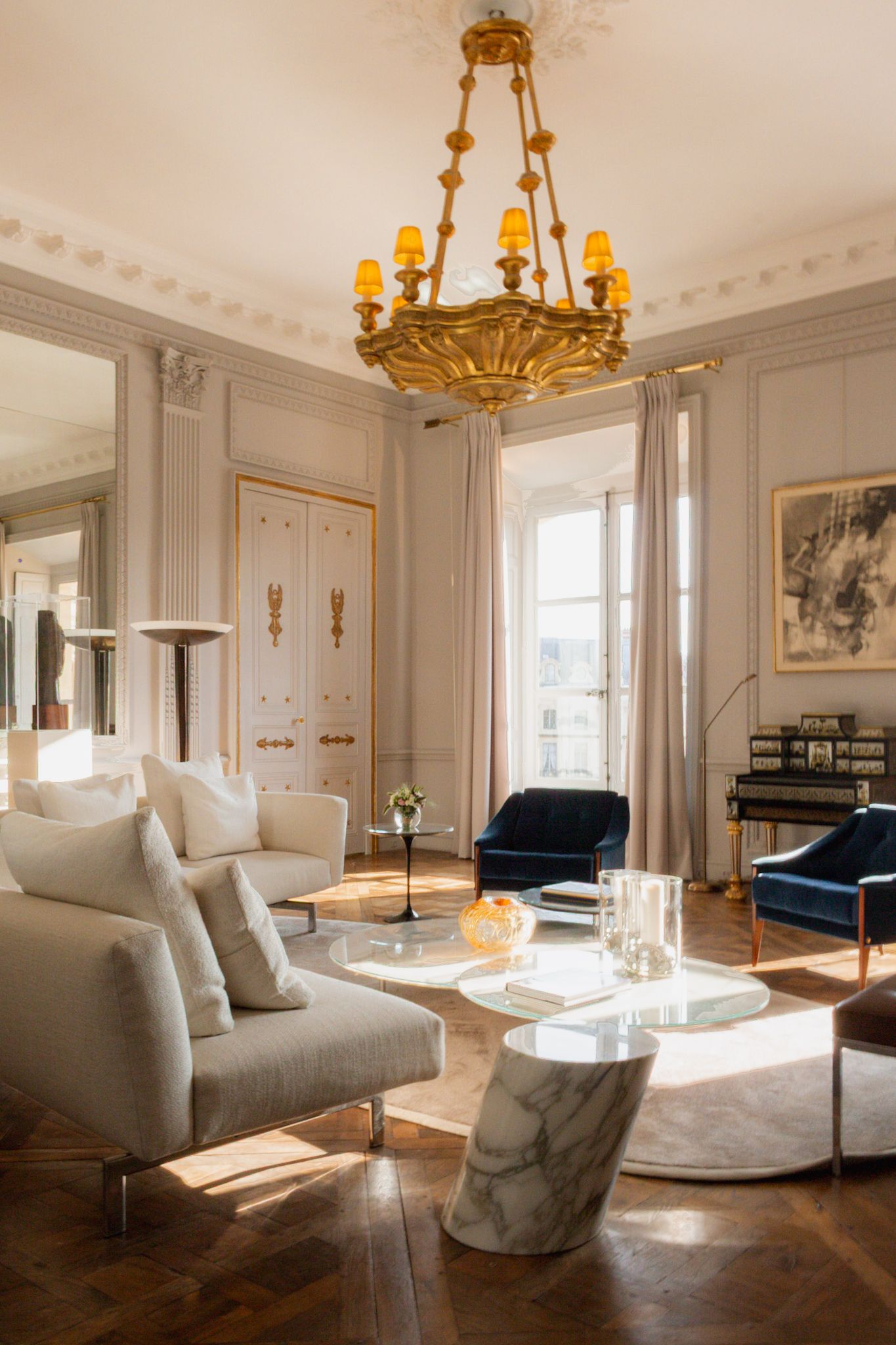 Luxurious Parisian-style living room with gold chandeliers, white modern furniture, and elegant crown molding in natural light.
