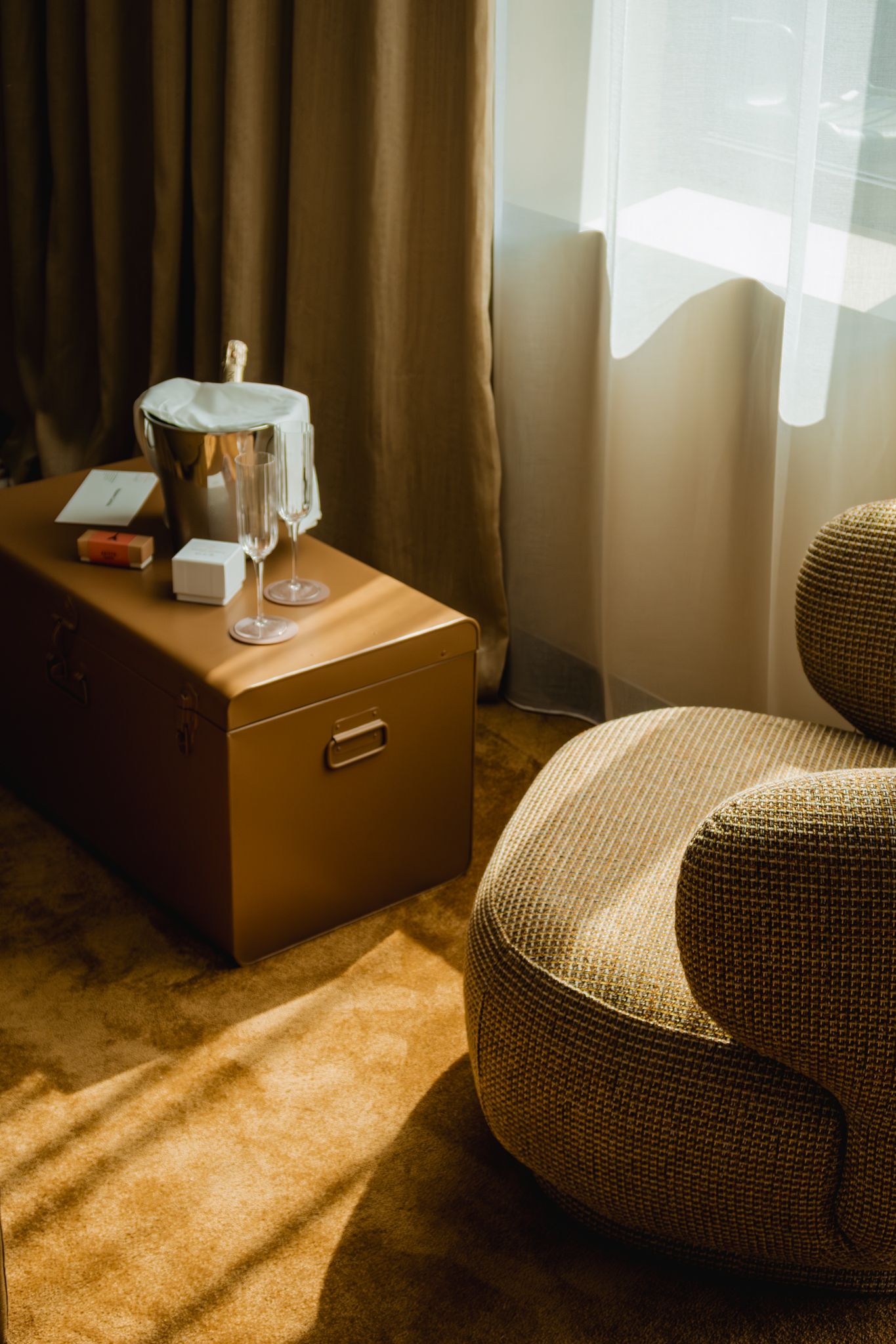 A cozy hotel room corner with an ice bucket on wooden cabinet, plush round chair and light filtering through curtains.