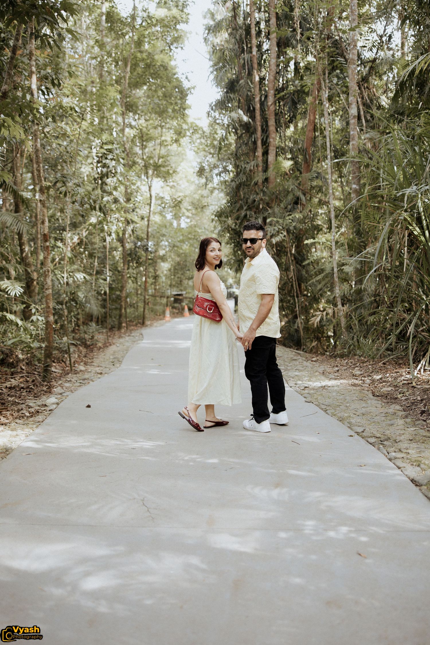 A couple poses on a concrete path surrounded by tall trees in a forest setting.