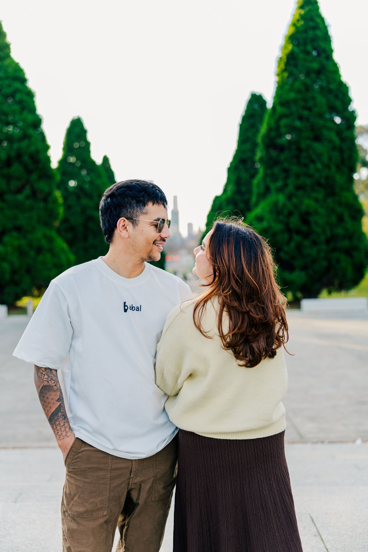 Couple in coordinated white and neutral outfits walking together near cypress trees.