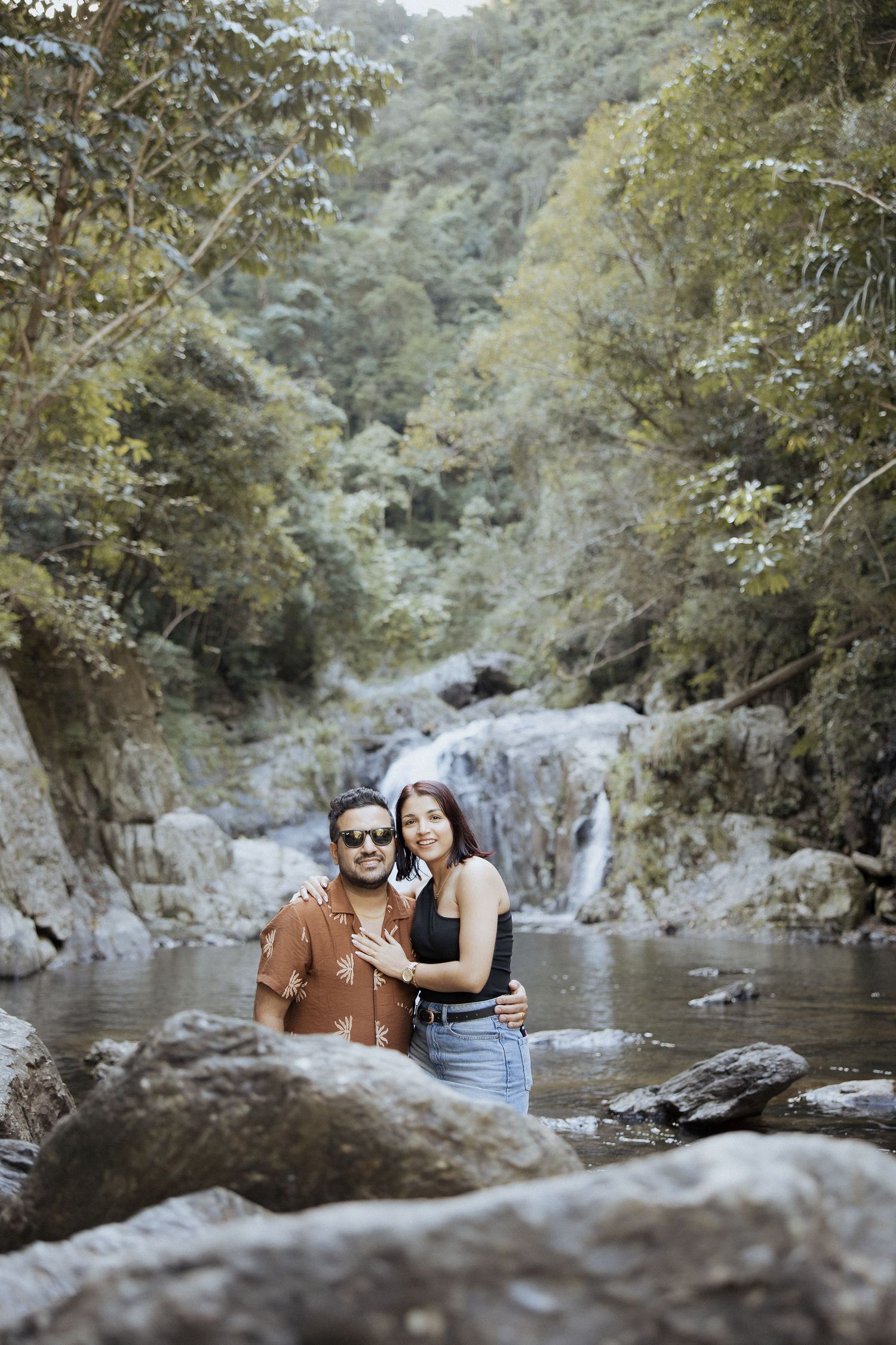 A couple poses in front of a scenic waterfall in a lush forest setting.