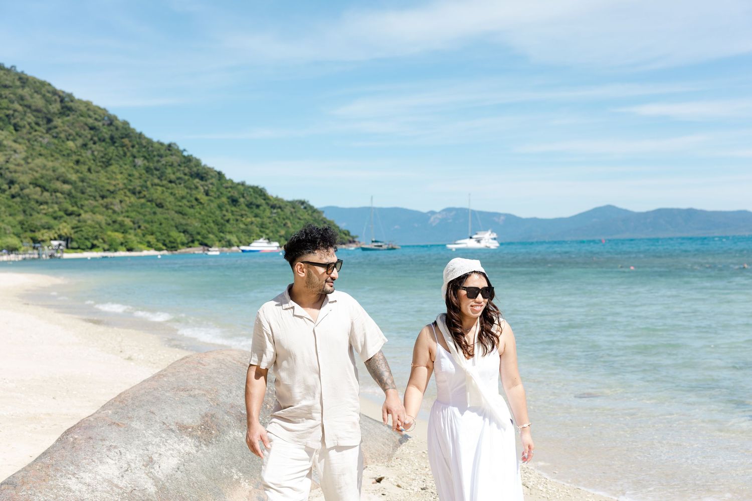 A couple strolls hand in hand along a pristine tropical beach with turquoise waters and mountains in the background.