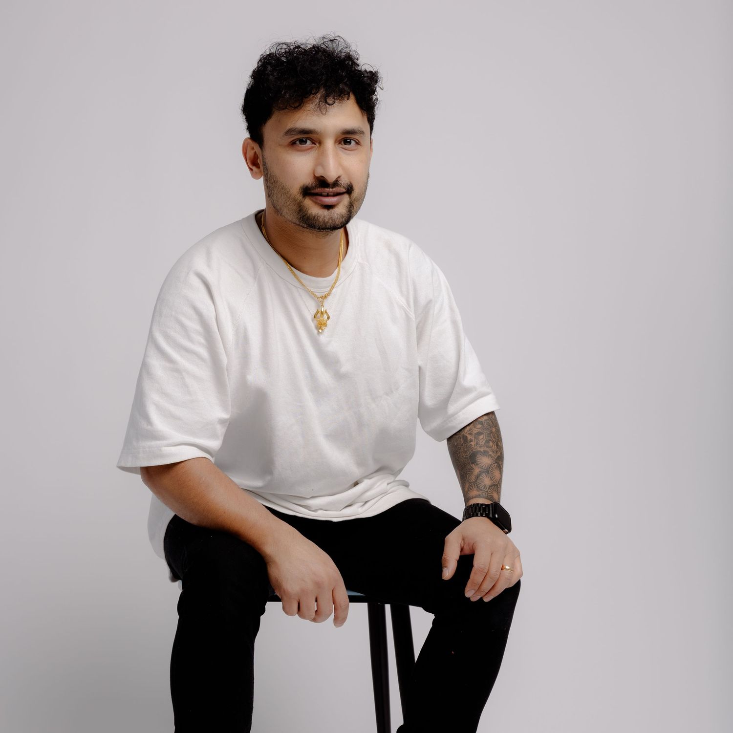 Person in white t-shirt and black pants sitting on dark stool against light gray background.