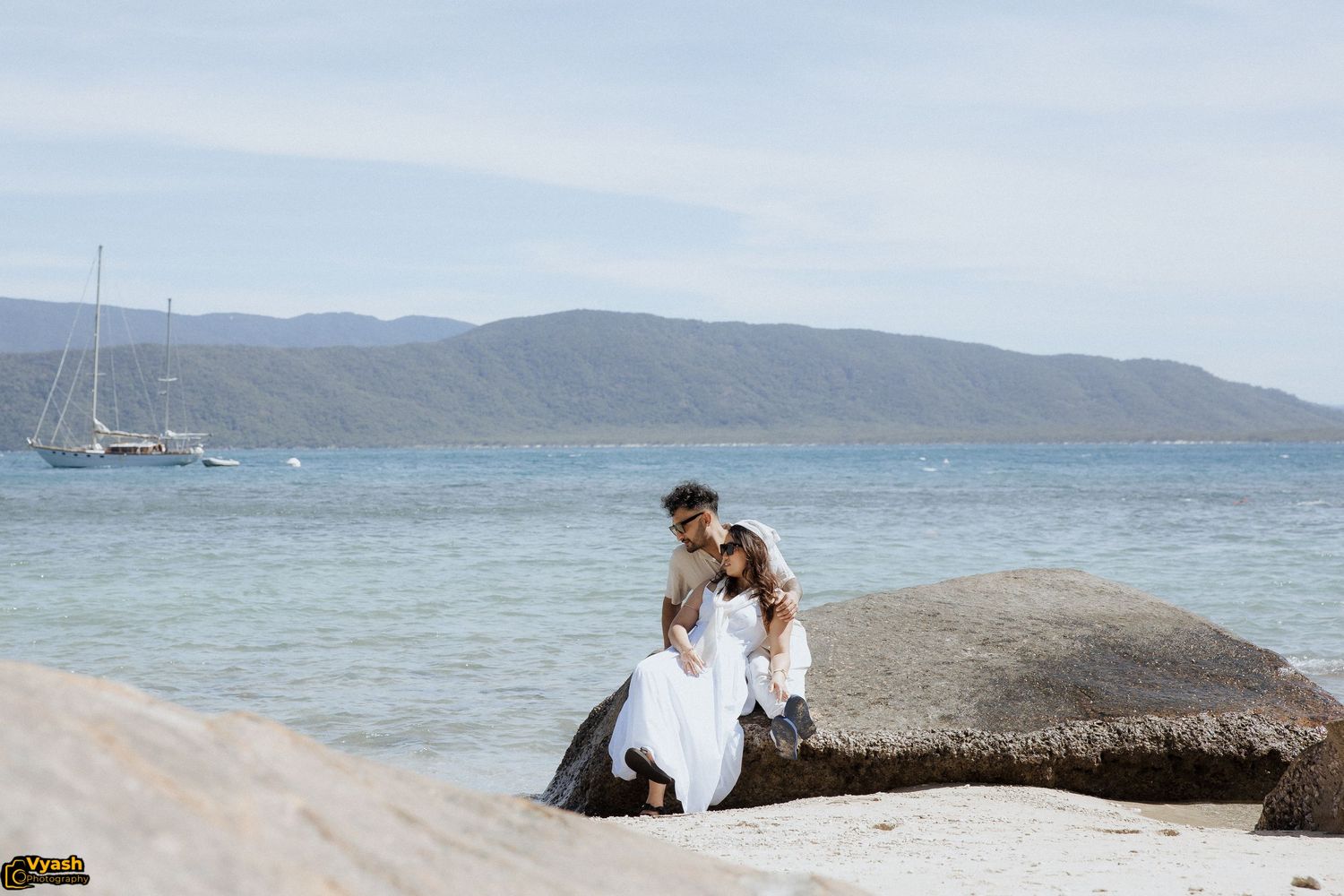 A couple sits together on coastal rocks with sailboats and mountains visible in the background.