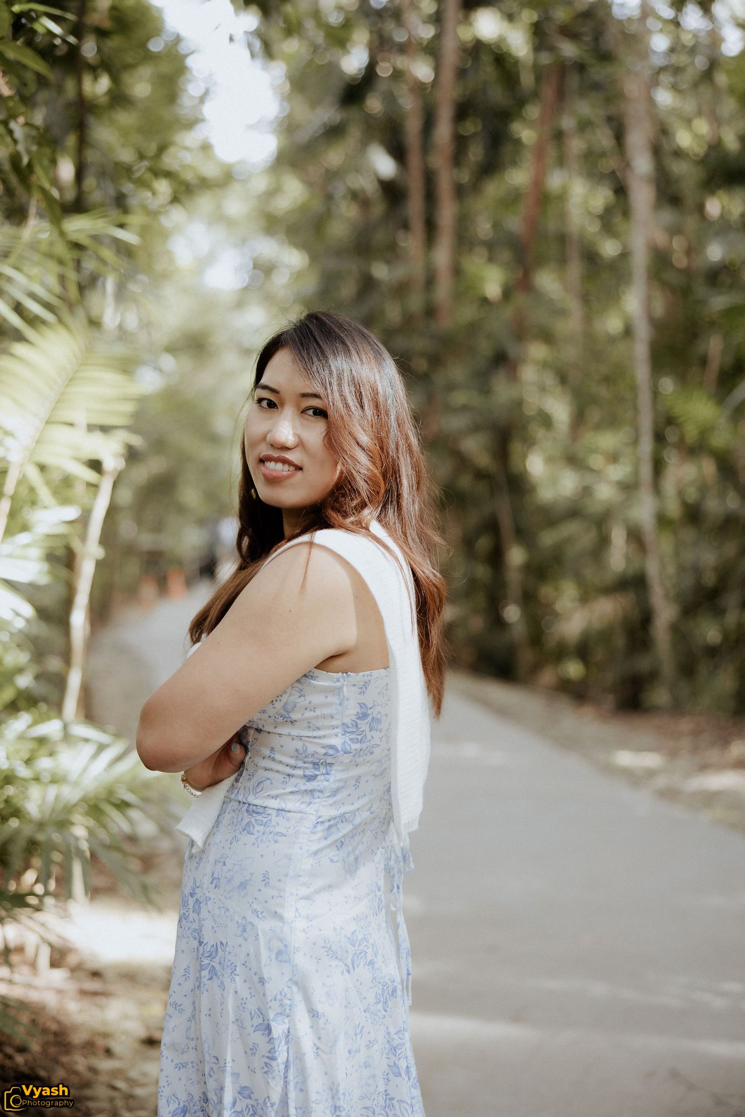 Person in a light blue floral dress poses on a tree-lined path in a natural outdoor setting.