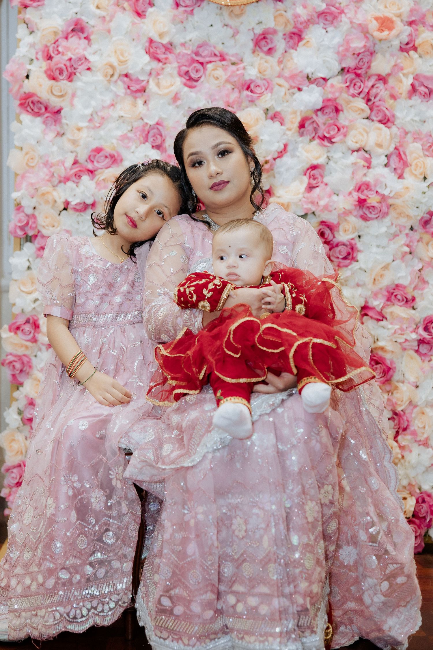 Three people in formal pink attire pose against a rose-covered wall backdrop for a special occasion.