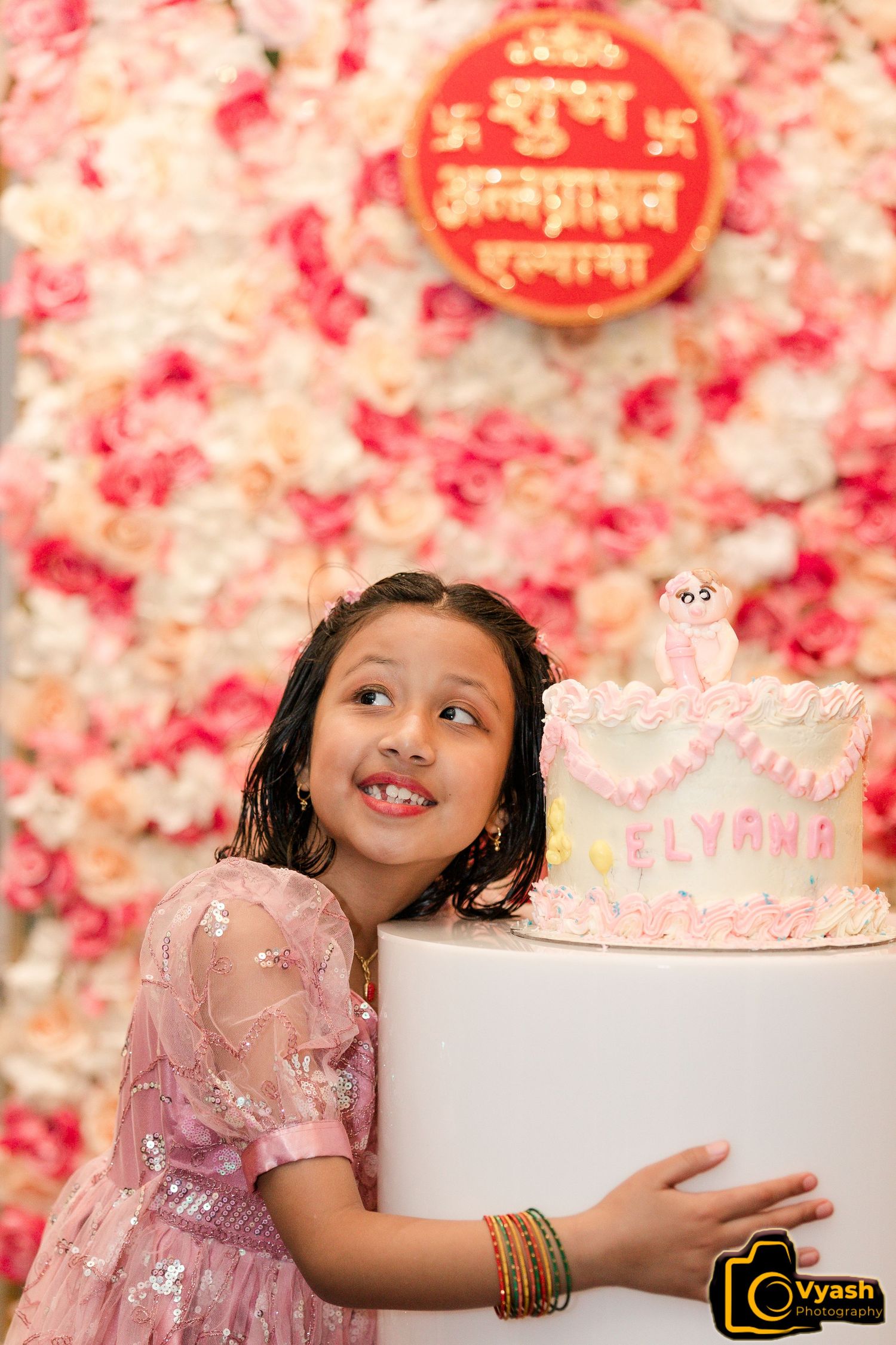 Pink birthday cake displayed against floral wall with celebration banner.
