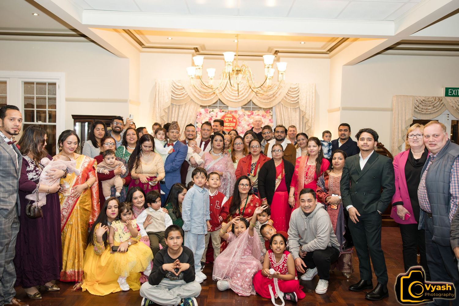 Large group photo of celebration attendees in traditional attire at an indoor venue.