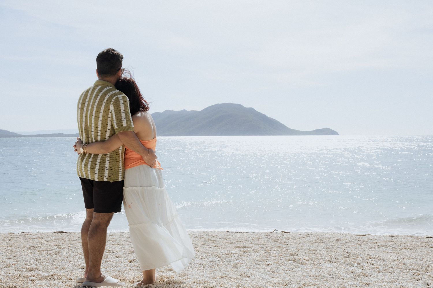 A couple embraces while looking out at the ocean and distant island from a sandy beach.