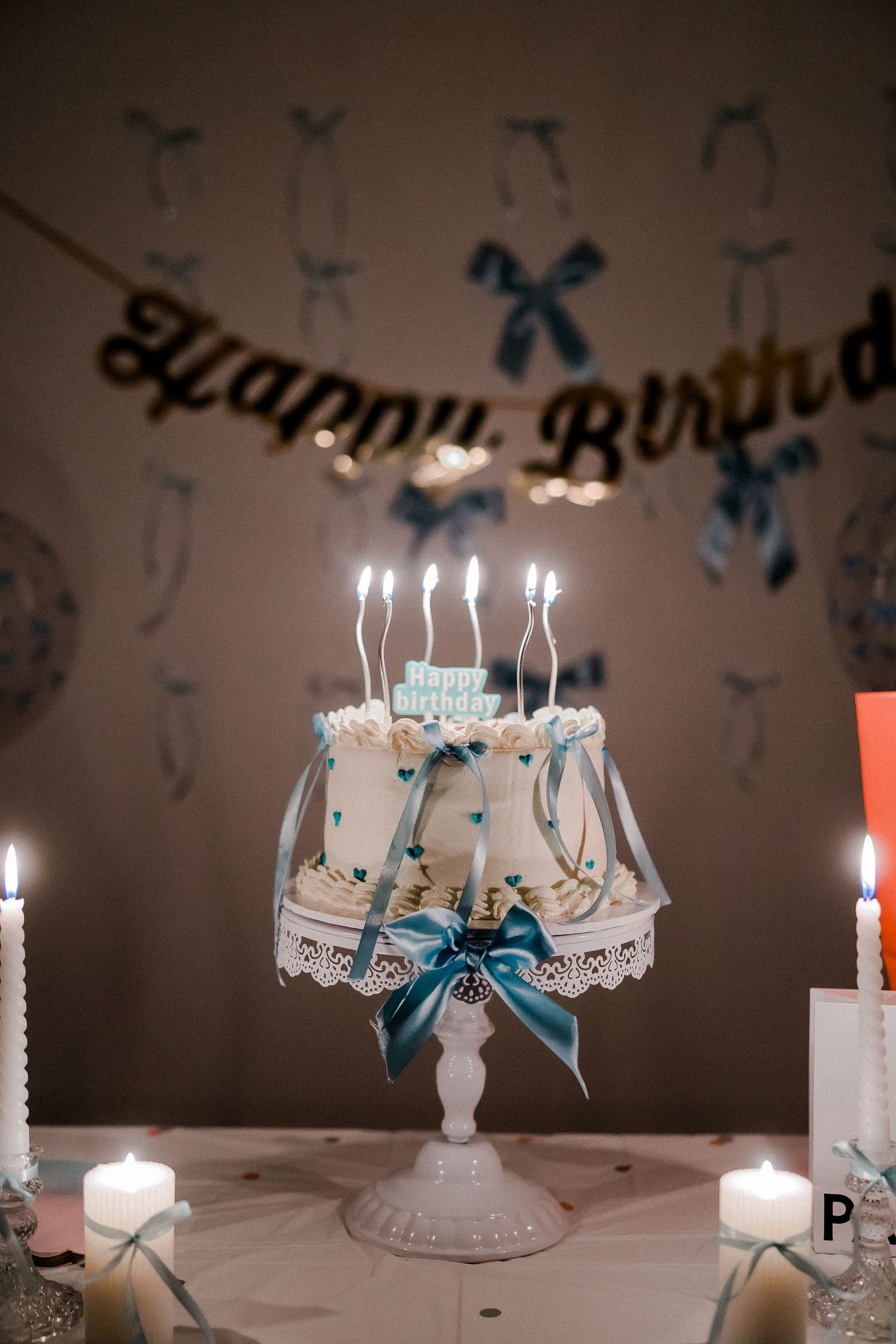 White birthday cake with blue ribbons and lit candles under a Happy Birthday banner.