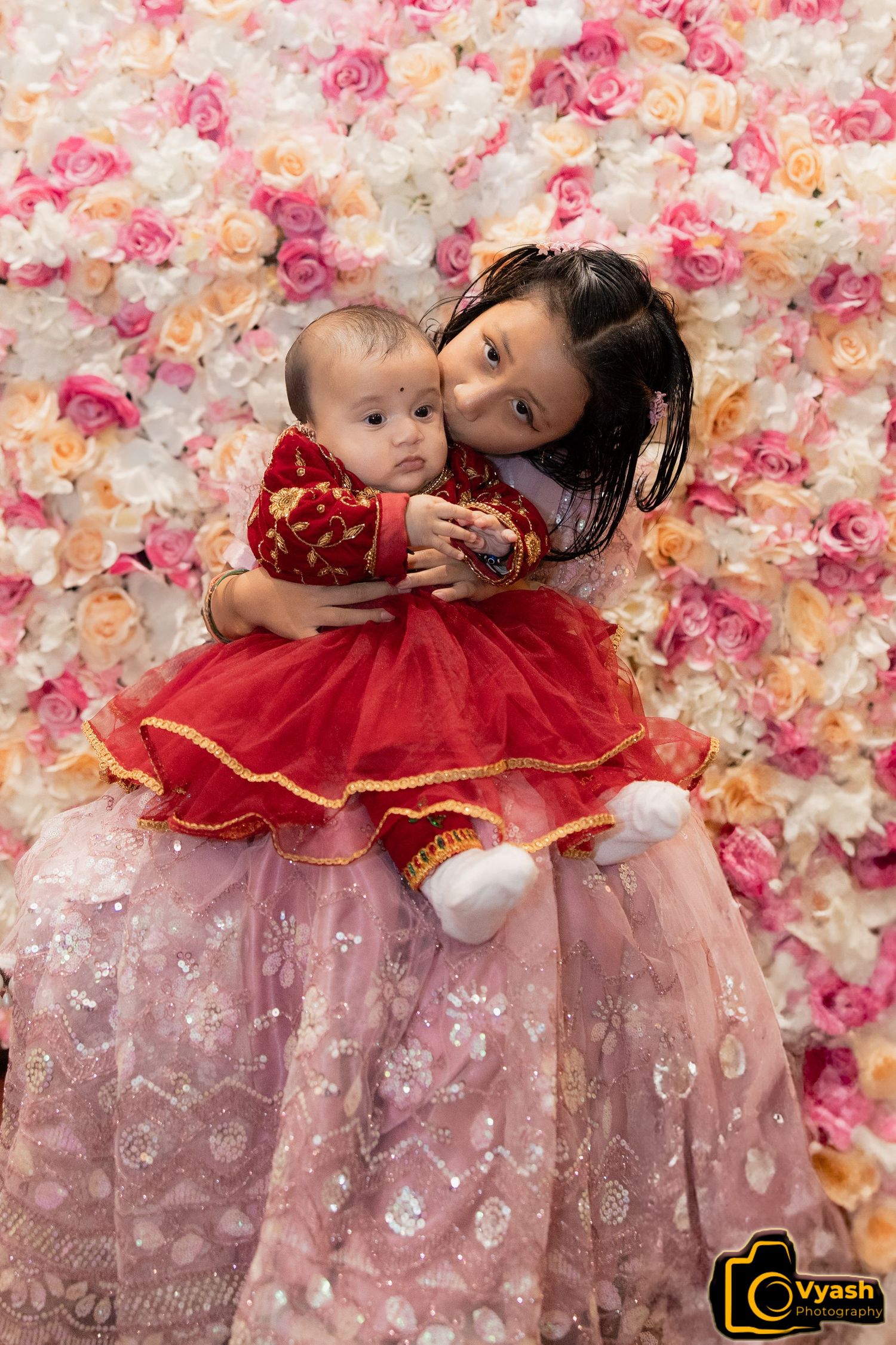 A tender moment between family members against a pink floral backdrop wearing traditional red and pink dresses.