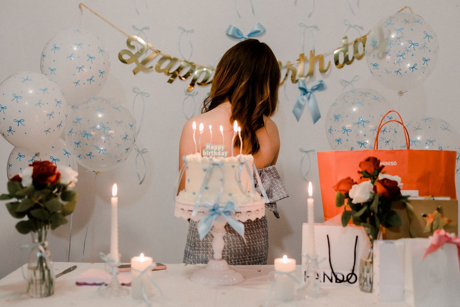 Birthday party setup with balloons, candles and gift bags on a white table.