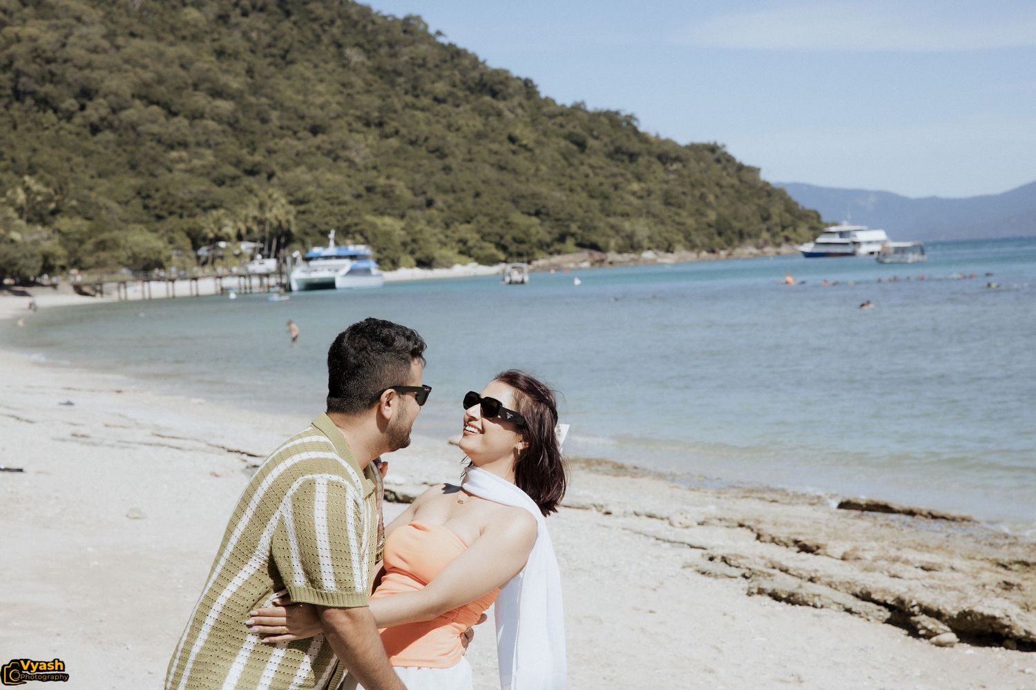 A couple in casual attire embraces on a scenic beach with boats and mountains visible in the background.