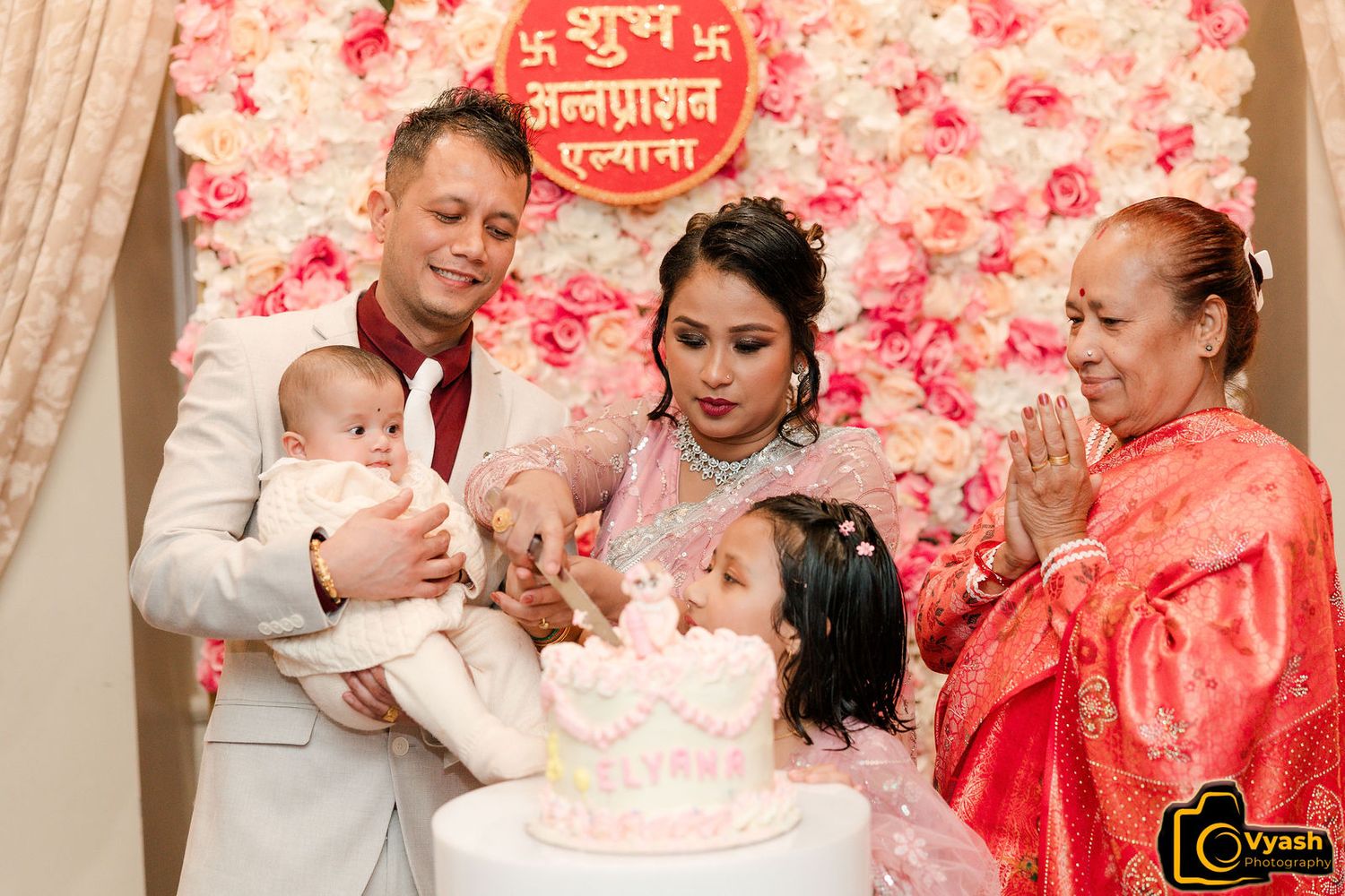 A family gathering around a pink birthday cake with floral backdrop for celebration.