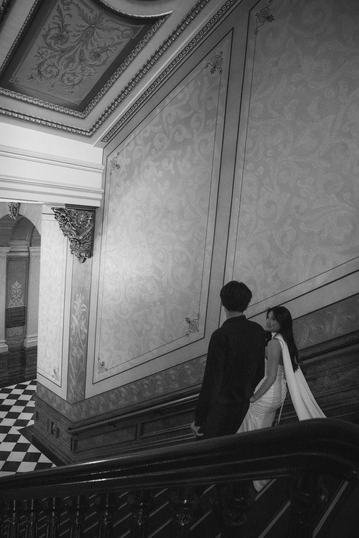 Black and white photo of a couple in wedding attire ascending an ornate staircase in an elegant historic building.