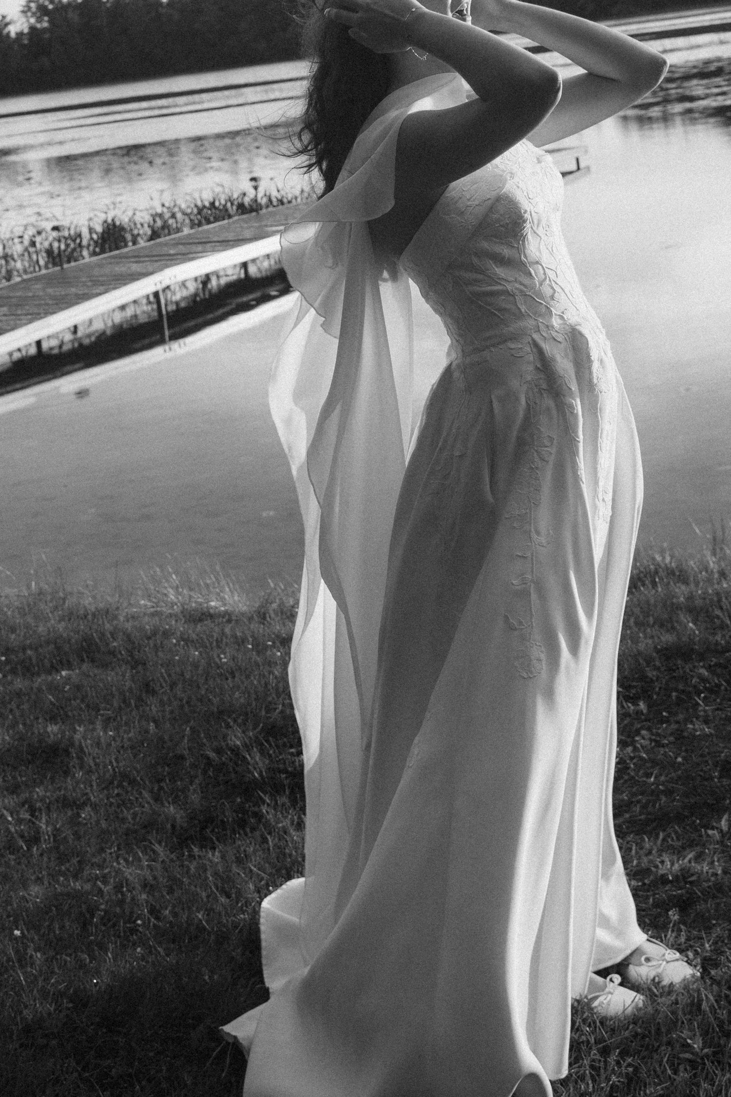 Black and white photo of a bride in a flowing white gown standing gracefully near a reflective pond.