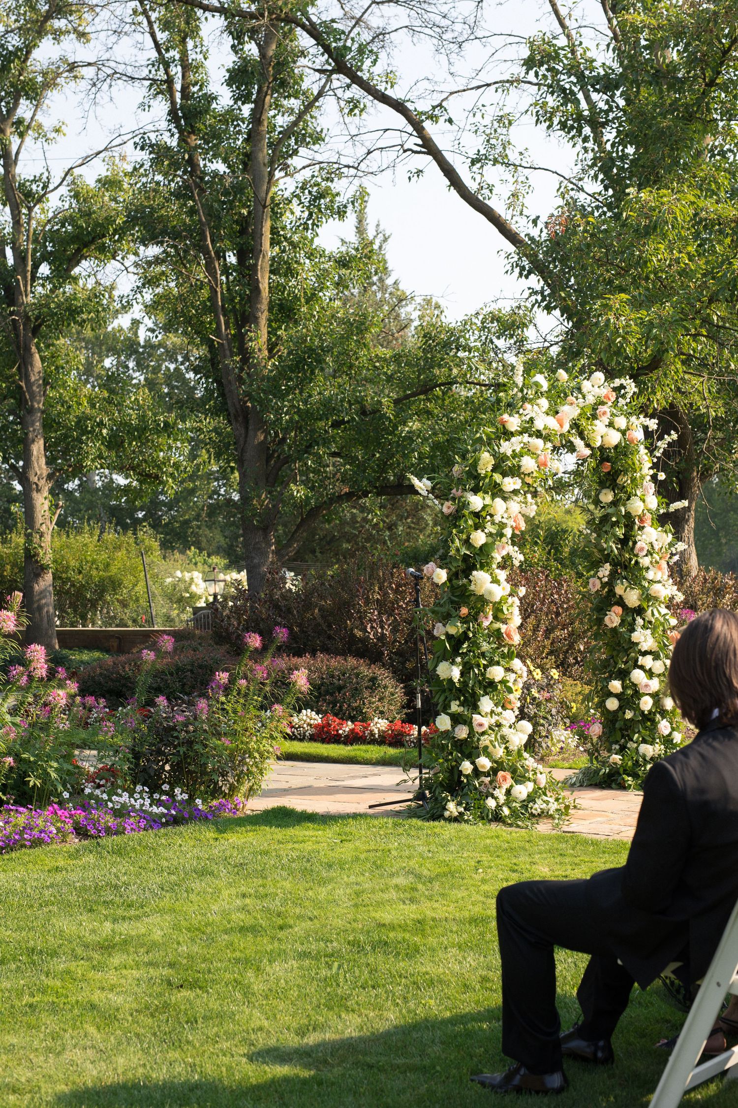 A garden wedding ceremony setup with white floral arches and pink blooms along a lush green lawn.