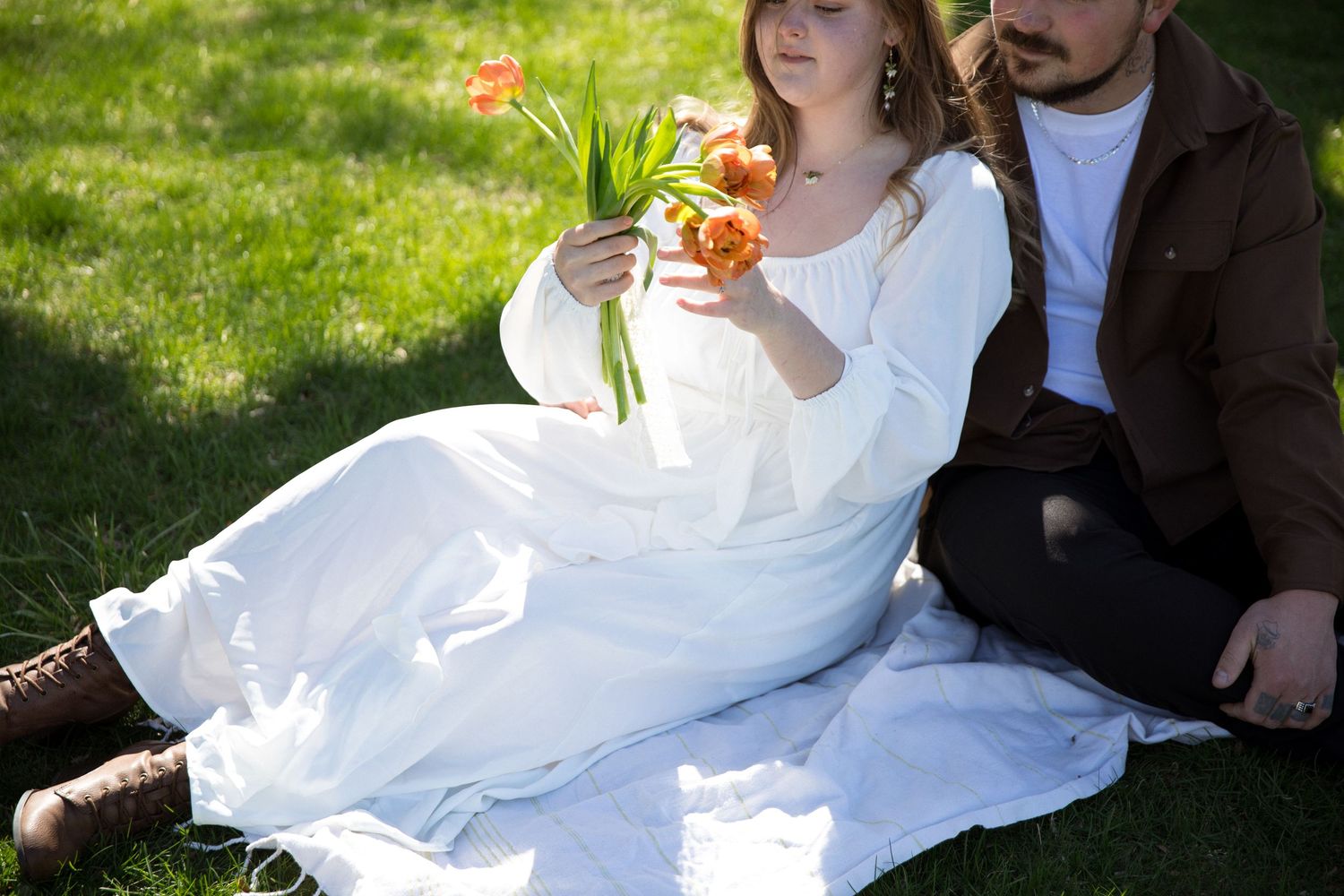 Couple sitting on white blanket in grass, woman in white dress holding orange tulips bouquet outdoors.
